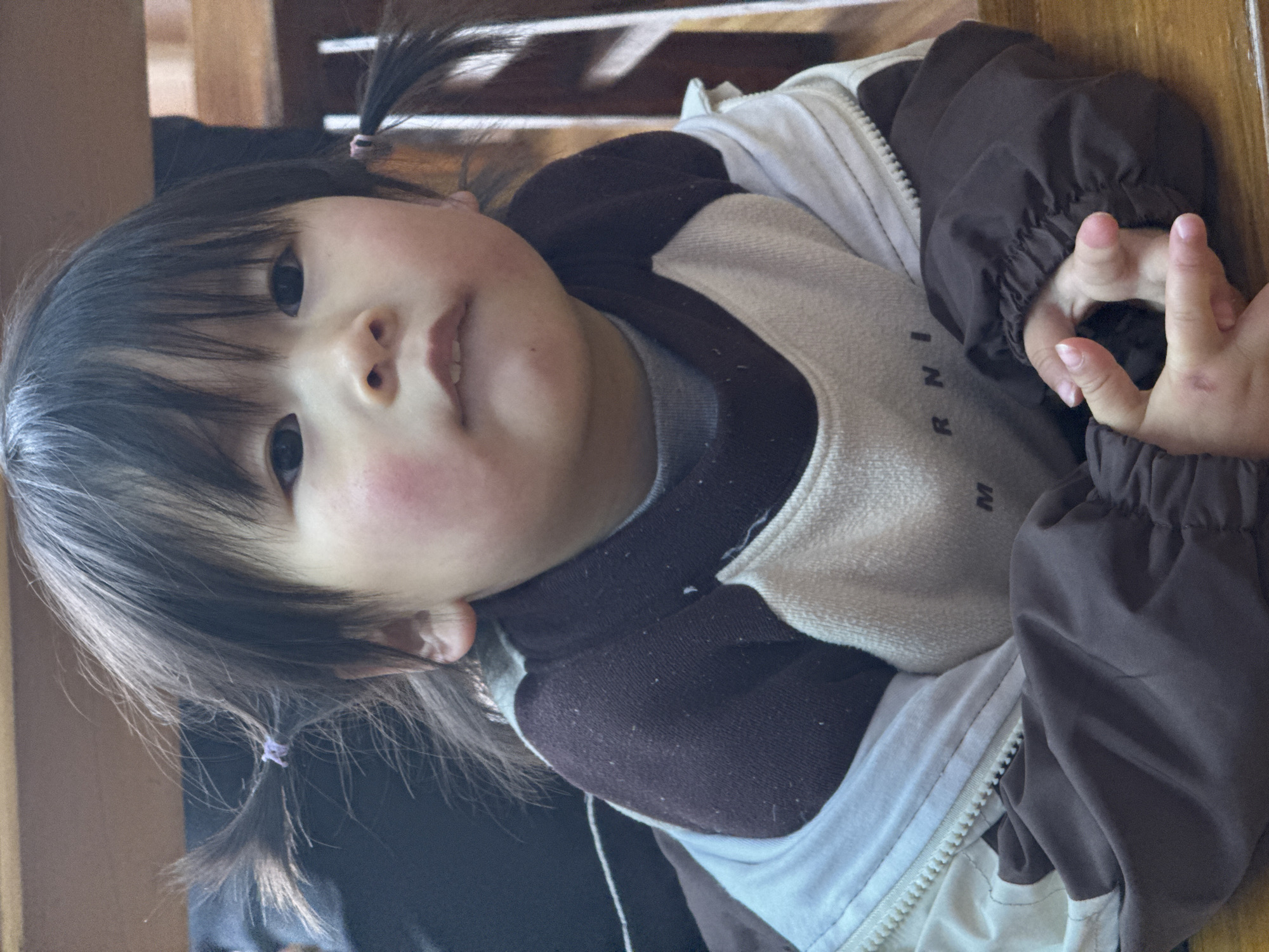 A young child with dark hair in pigtails is looking at the camera. The child is wearing a cream and brown zip-up jacket over a gray sweatshirt. The background appears to be a wooden chair or table. The location is presumed to be Qianjiang, China.