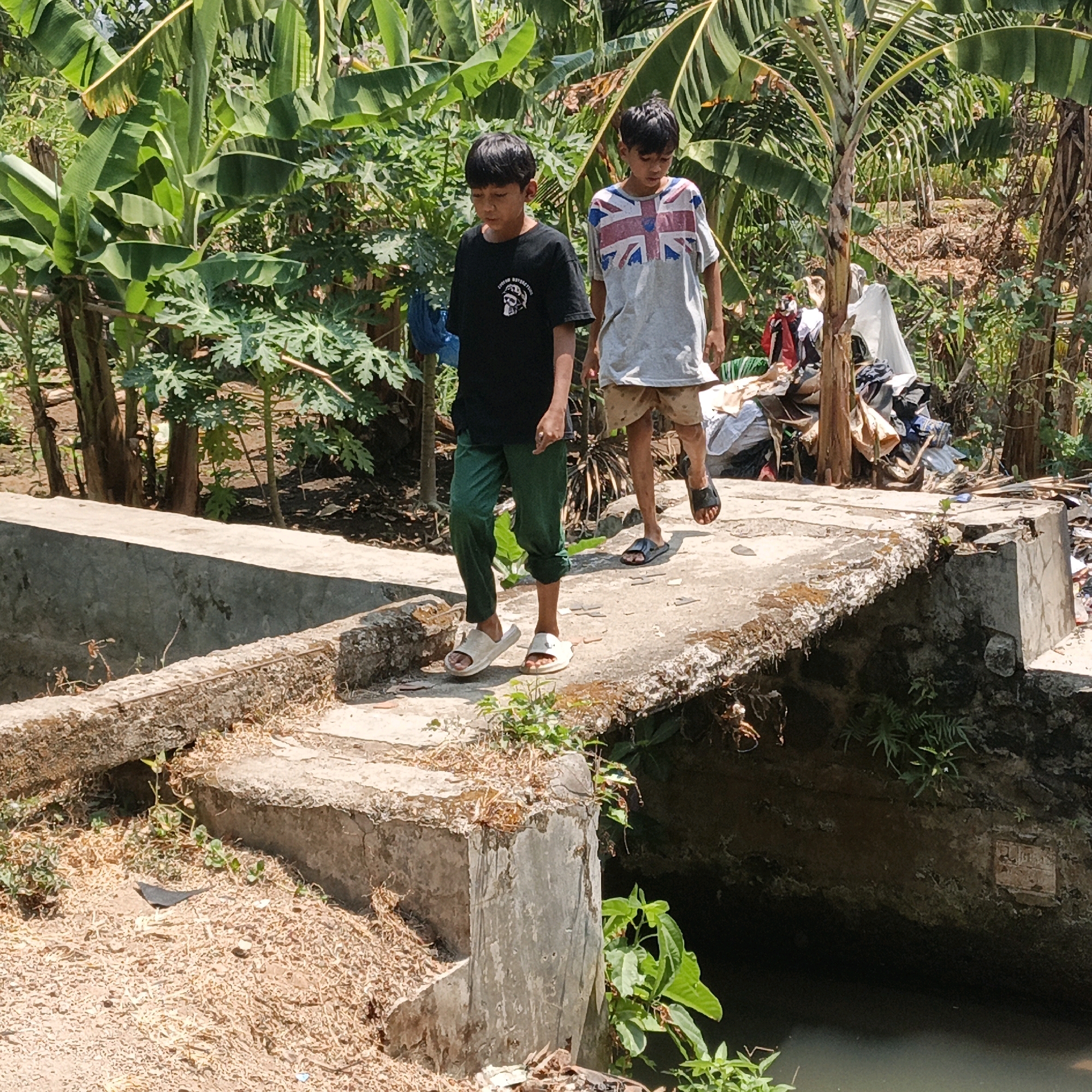 Two young boys are walking across a concrete bridge in Mangli, Indonesia. The boy on the left is wearing a black t-shirt and green pants, and the boy on the right is wearing a grey t-shirt with a Union Jack design and light brown shorts. They are walking away from the camera, towards a pile of laundry and debris on the far side of the bridge. The bridge spans a small body of water, and the surrounding area is lush with banana trees and other vegetation. The sky is bright and sunny, indicating it is daytime.