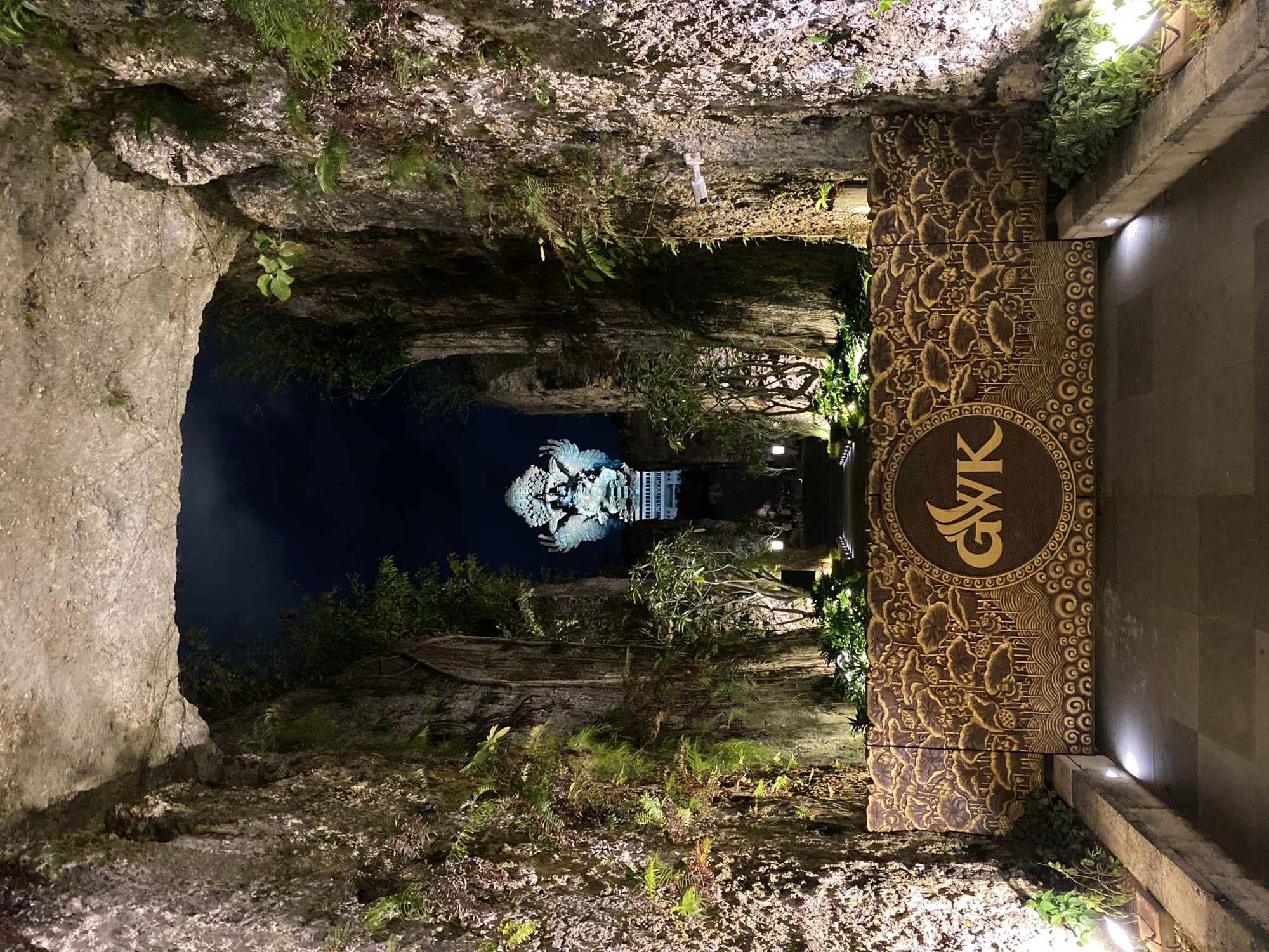 The image captures the GWK Cultural Park in Kuta Selatan, Indonesia, at night. The foreground features a pathway leading through a rock formation, with lush greenery and artificial lighting illuminating the textured stone walls on either side. A decorative wall with an intricate lotus pattern and the "GWK" logo is prominently displayed. In the distance, a large statue of a deity, likely Garuda Wisnu Kencana, is illuminated against the dark night sky, standing atop a modern structure. The overall scene conveys a sense of grandeur and cultural significance, enhanced by the dramatic lighting and the natural rock environment.