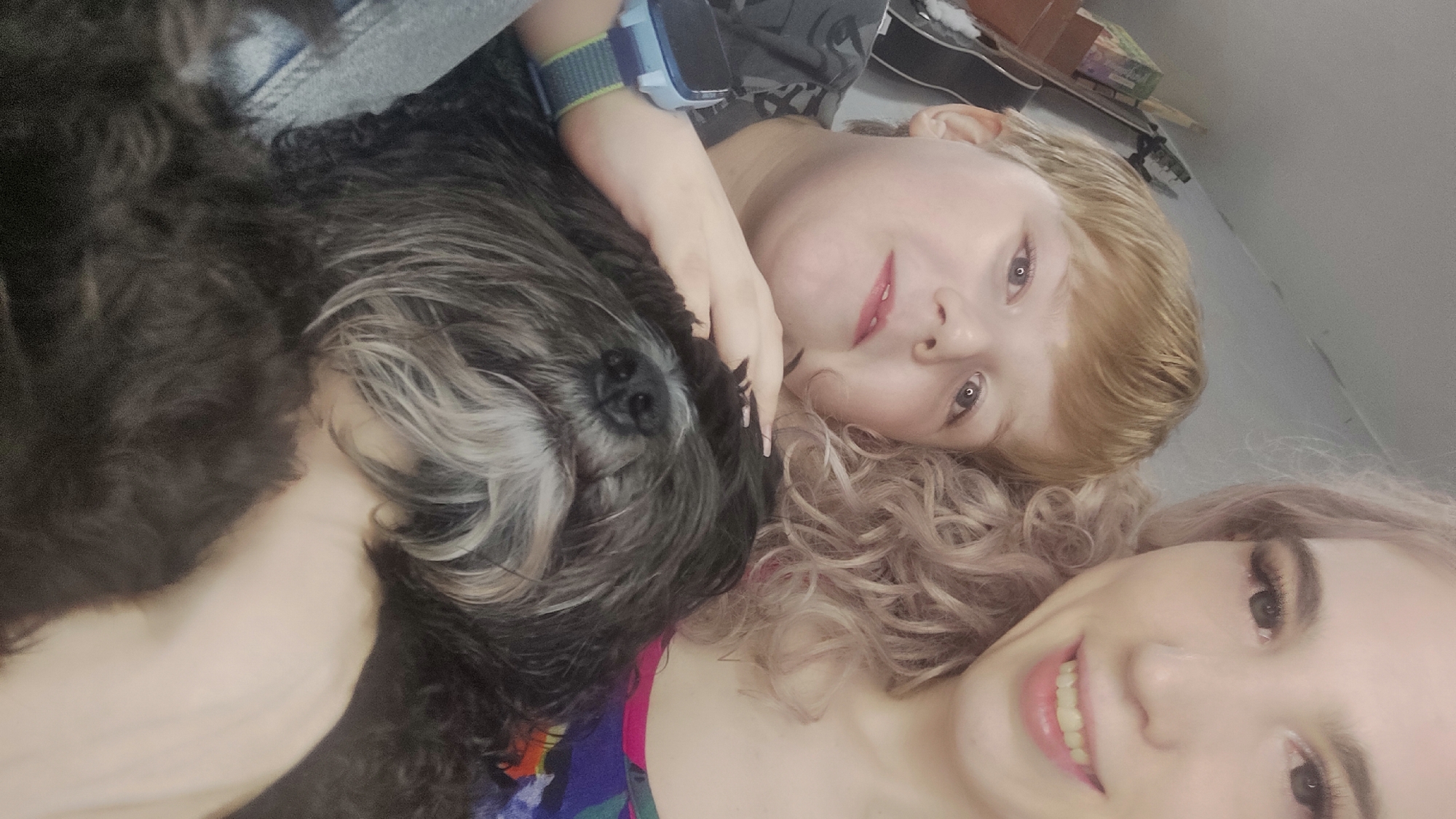 A close-up shot depicts two people and a dog in an indoor setting. One individual has light blond hair, light skin, and is wearing a grey patterned shirt and a blue smartwatch on their left wrist. Their face is oriented towards the viewer, resting near a black and grey dog, with their hand near the dog's head. The dog is lying down, with its face and body visible. A second individual, with light skin and curly pink hair, is positioned closer to the camera and is smiling. A portion of a bright, multicolored floral shirt is visible on their torso. The background includes a glimpse of a wooden object and a box. It is impossible to determine if the photo was taken in the city of Chaikovsky, Russia, based on the provided details.