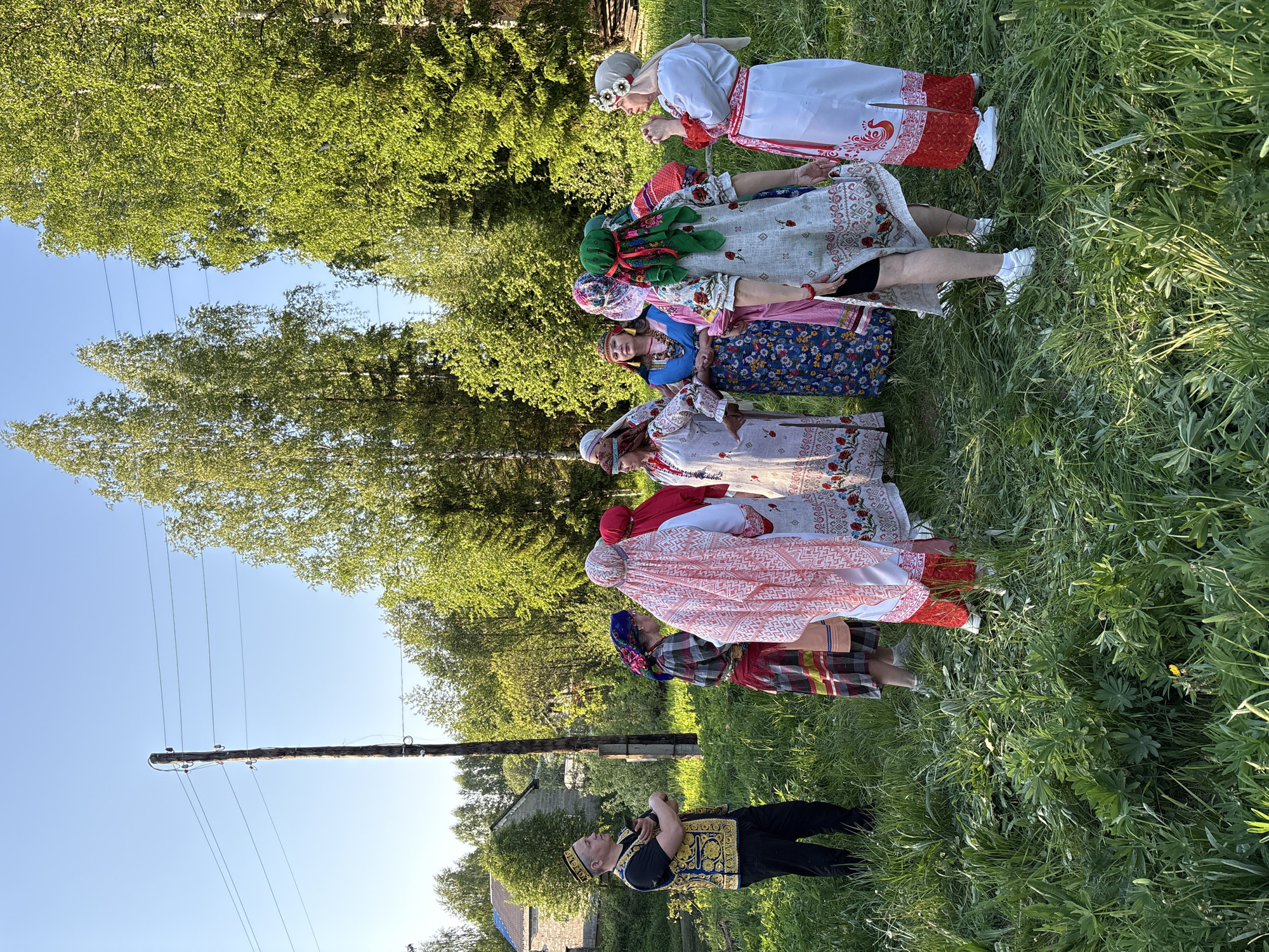 The image depicts a group of people in traditional Russian folk costumes standing in a grassy field in Markovo, Russia. From bottom to top, the first person is a man dressed in a black shirt and pants with a gold embroidered vest and hat. He is standing with his arms crossed, possibly observing the women in front of him. The next six people are women wearing traditional Russian dresses, skirts, and head coverings of various colors and patterns, white sneakers. They appear to be holding hands or touching each other in a line. The background features a wooden utility pole with wires, green trees, and the clear blue sky. Also a house is partially visible. The scene suggests a cultural performance or celebration taking place outdoors.