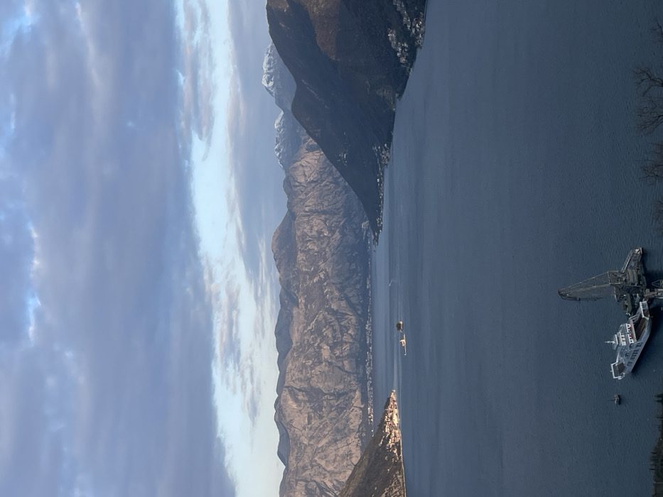 An elevated, wide-angle forensic image depicts the Bay of Kotor in Montenegro, specifically observed from the hills above Morinj, looking southeast towards the inner bay. The sky above is partially overcast with dark grey and lighter blue stratocumulus clouds. In the background, a substantial range of rugged, grey-brown mountains stretches across the horizon, exhibiting visible geological features and deep shadows; some distant peaks on the far right are capped with snow. The midground is dominated by the calm, dark blue waters of the bay.
Along the left shoreline in the mid-distance, a dense settlement, identifiable as Perast, features numerous buildings with red roofs. In the center-left of the bay, two distinct islands are visible: the artificial island of Our Lady of the Rocks, characterized by its prominent white church and surrounding structures, and the smaller, naturally vegetated island of Sveti Đorđe slightly to its right. The right shoreline shows a scattering of residential buildings extending into the mid-distance.
In the immediate foreground, positioned in the water, a white and blue vehicle ferry is moored next to a large dark grey floating crane or dredge rig with a prominent boom. A small, dark cylindrical buoy is situated near these vessels. Sparse, dark tree branches are visible in the extreme bottom-right corner, indicating the elevated vantage point. No human figures or discernible activity are evident in the scene.