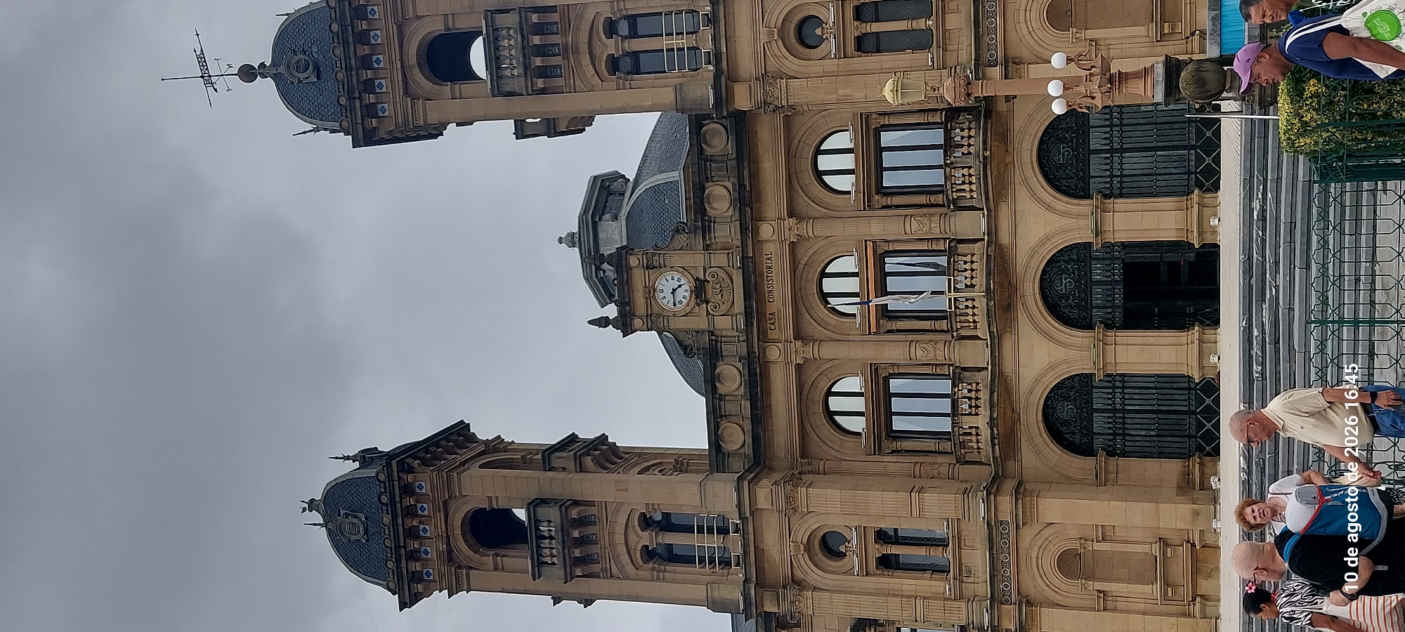 The image shows a view of the Casa Consistorial in San Sebastián, Spain, a large, ornate building with a stone facade, two prominent towers, and a central dome. A clock is visible on the front of the building, and a weather vane is mounted on one of the towers. The sky is overcast. In the foreground, several people are visible, including men and women of various ages, some of whom are looking towards the building, and others appear to be walking past. One person is carrying a white tote bag with a green design. A timestamp overlay at the bottom left of the image reads "10 de agosto de 2026 16:45". The architectural style is ornate and appears to be from the late 19th or early 20th century. The overall scene suggests a public square or plaza in front of the city hall.