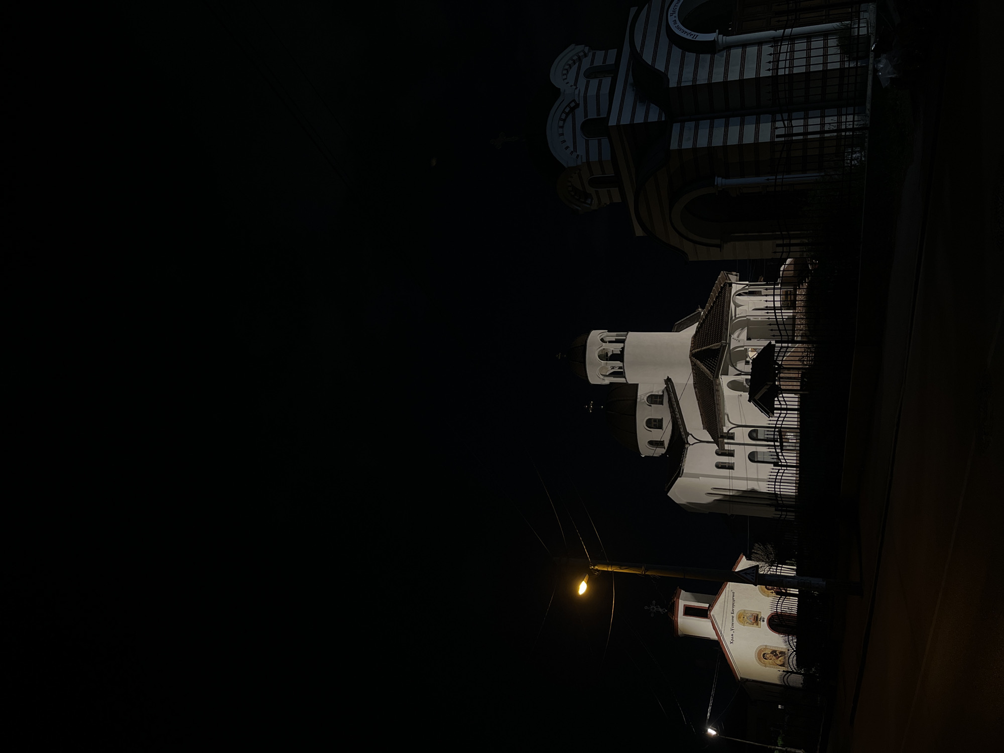 The image depicts a nighttime scene on a street in Sofia, Bulgaria. Three buildings are visible, presumably churches, illuminated against the dark sky. The closest building is small with a red roof and painted icons on the facade. A street light is visible in front. A mid-sized building with domes is set back from the street, and a large, white church with an arched facade is positioned farther in the background.