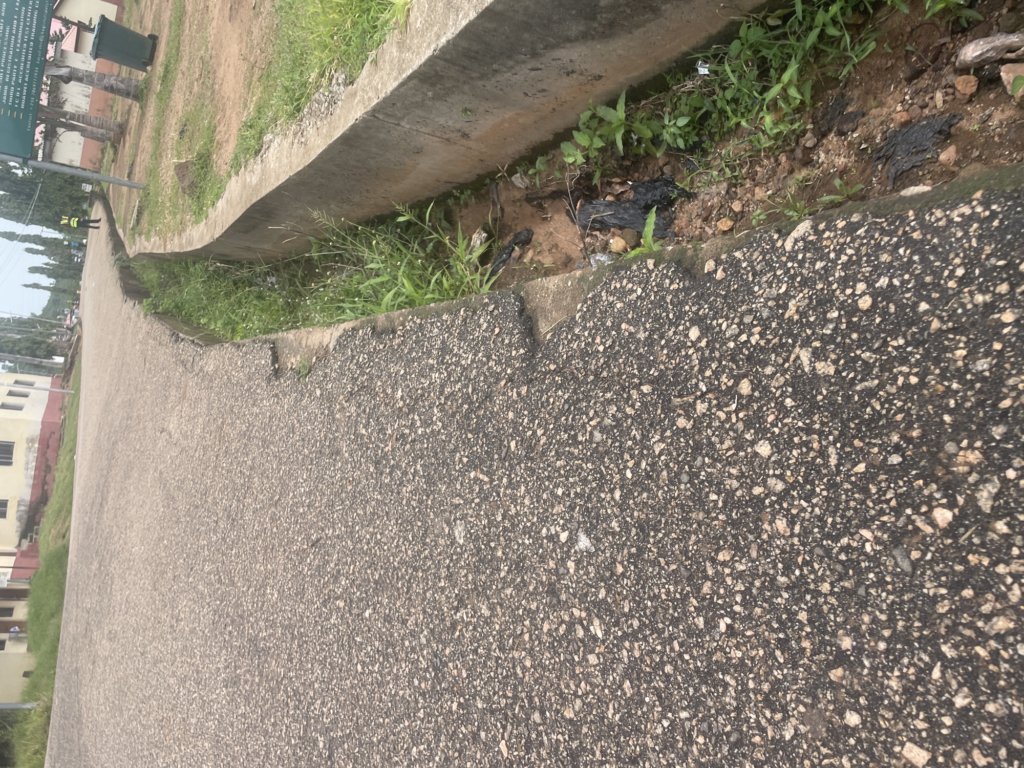 The image depicts a paved road with a concrete drainage channel running alongside it. The road surface is asphalt with visible aggregate. The drainage channel is partially filled with soil, debris, and some black plastic refuse. Green vegetation, including grasses and small plants, grows within and around the drainage channel. In the background, a building with a red trim is visible, along with several tall trees. Further down the road, a person in a high-visibility yellow vest stands near a street sign. A green refuse bin is also present in the background. The overall scene suggests an outdoor environment in an urban or semi-urban area, possibly a roadside within Lokoja, Nigeria, characterized by the presence of infrastructure and vegetation.