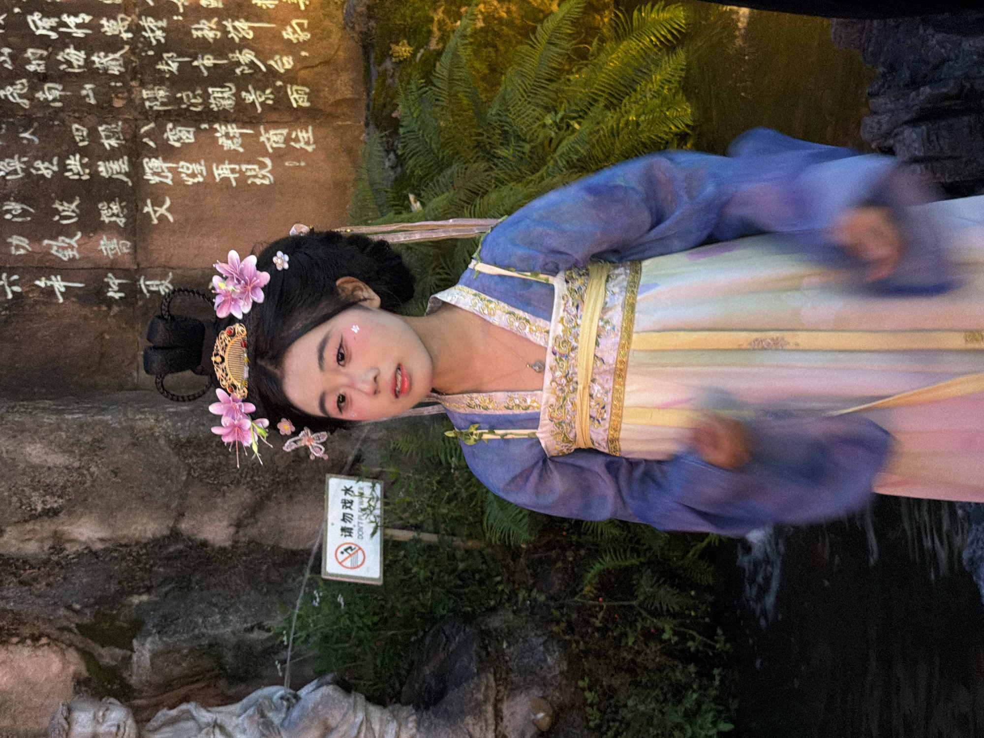 This media file shows a young woman dressed in traditional Chinese Hanfu attire, possibly posing for a photograph. She has her hair styled in an updo adorned with pink and white flowers and a decorative comb. Her makeup is subtle, with a touch of pink blush on her cheeks and eyes. The setting appears to be outdoors, near a rock face with inscribed calligraphy. Lush green ferns and foliage are visible, suggesting a garden or natural environment. A small white sign with red lettering and a prohibition symbol is also present, reading "请勿戏水" (Do not play in the water) and "DON'T P". The overall mood is serene, with the woman's expression conveying a gentle, almost ethereal quality. The lighting suggests it might be evening or a dimly lit area. The presence of water, possibly a fountain or stream, is hinted at by the blurred motion in the lower right portion of the image. The inscribed calligraphy on the rock face adds a historical and cultural dimension to the scene. The woman's dress is a flowing multi-colored garment with intricate golden embroidery on the bodice.