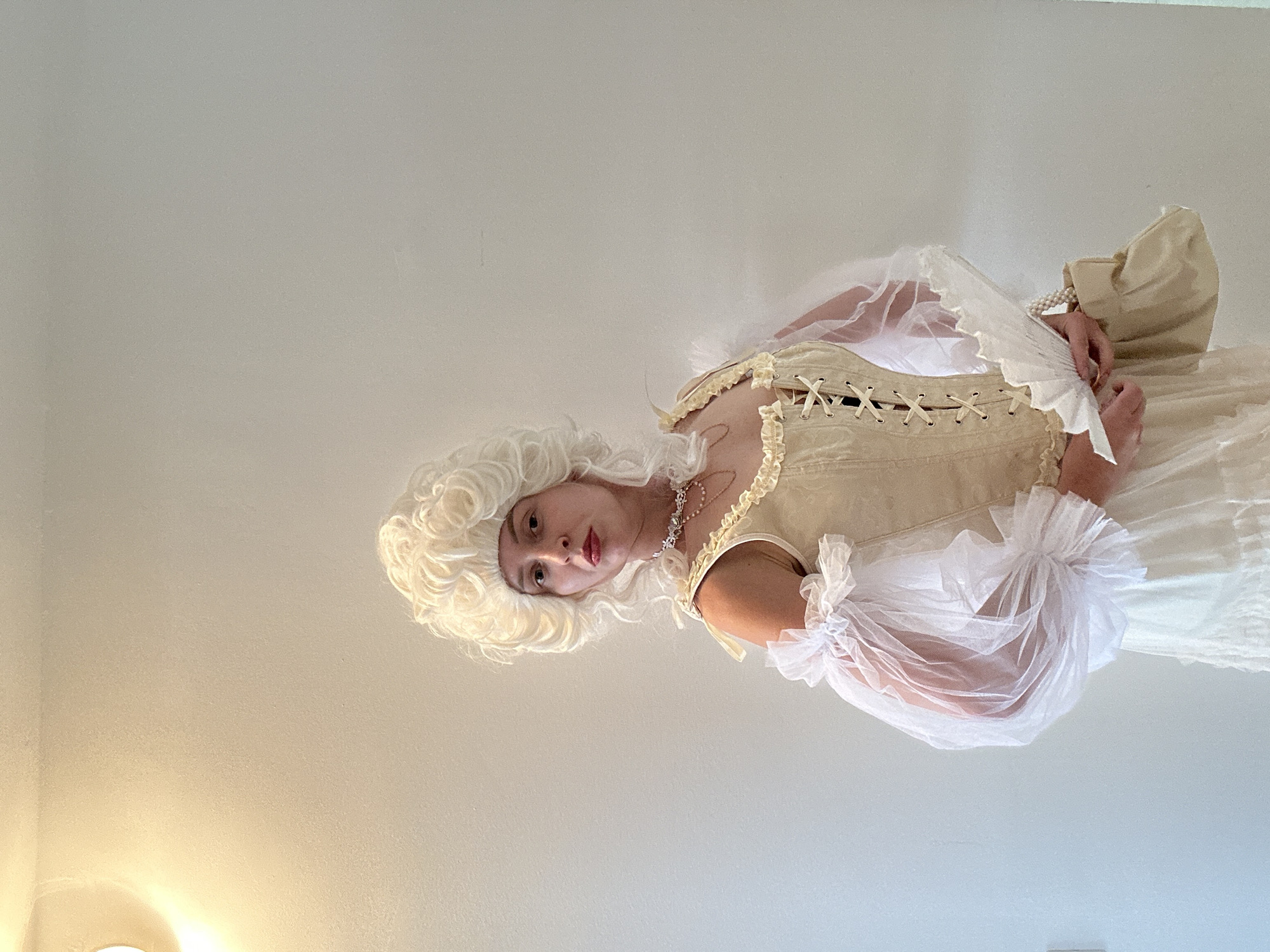 A person wearing a cream-colored, historical-style dress and a white wig stands against a plain, light-colored wall. The dress features a laced bodice, ruffled sheer sleeves, and a tiered skirt. The person holds a white fan and a small cream-colored bag. A pearl necklace is visible around their neck. The scene appears to be indoors, with a light fixture visible in the upper left corner. There is no indication of the city of Petah Tikva, Israel in the image.