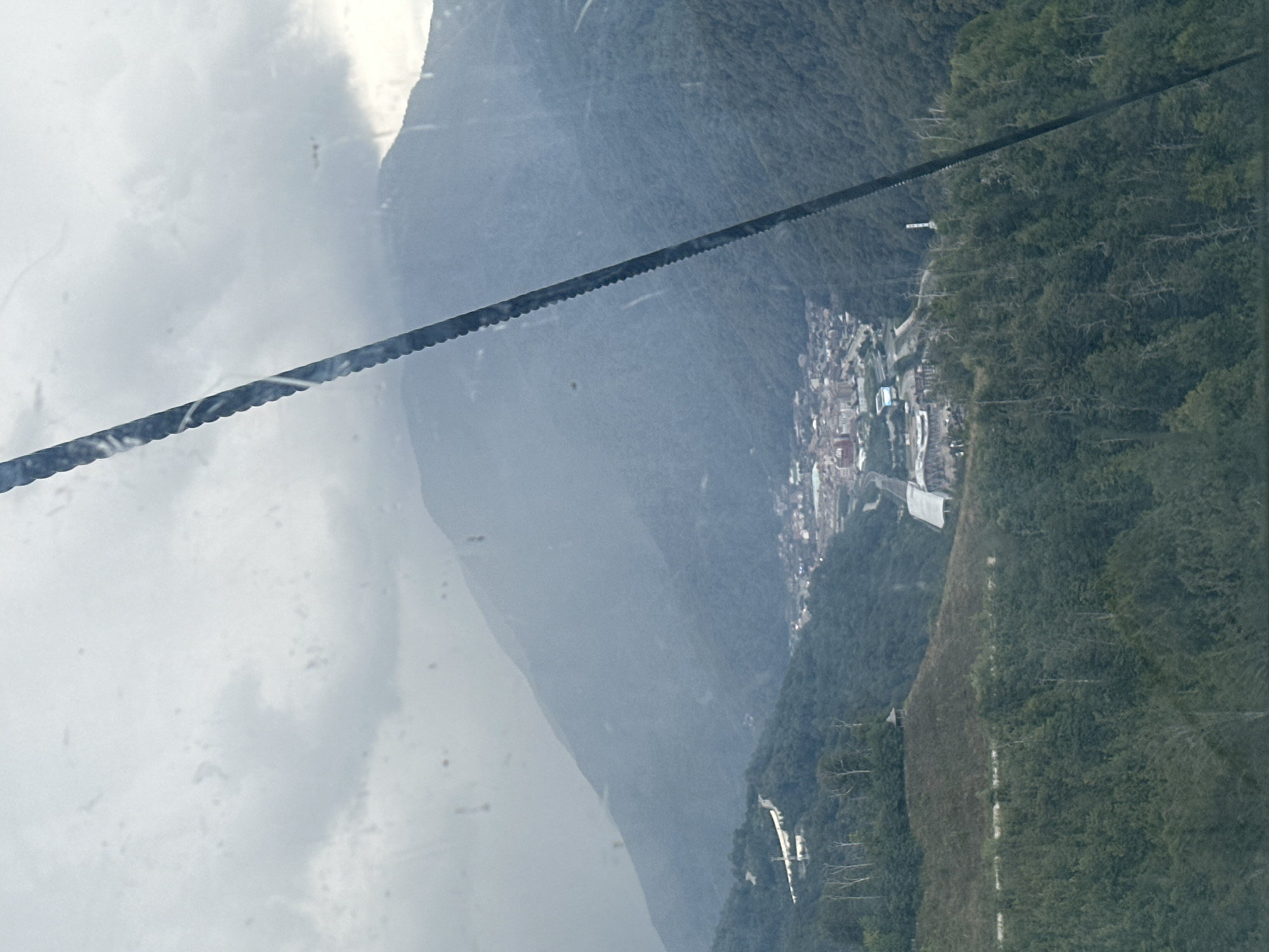 The image is a wide-angle, eye-level shot of a mountainous landscape seen through a window with some smudges or water droplets. A thick, dark, serrated cable stretches diagonally from the top right to the middle of the frame, partially obstructing the view. In the mid-ground, a small town or village, likely Estosadok, Russia, is nestled in a valley between dense green mountains. Buildings in the town appear to be clustered together, with some evidence of roads and infrastructure. The mountains surrounding the town are covered in thick, dark green forests, with some lighter patches indicating paths or clearings. The sky is overcast with heavy, gray clouds, and a hazy, atmospheric effect softens the distant mountain ranges. The foreground is dominated by more dense forestation on a steep slope, with several bare, spindly trees. The overall impression is one of a remote, natural setting with human habitation integrated into the valley.