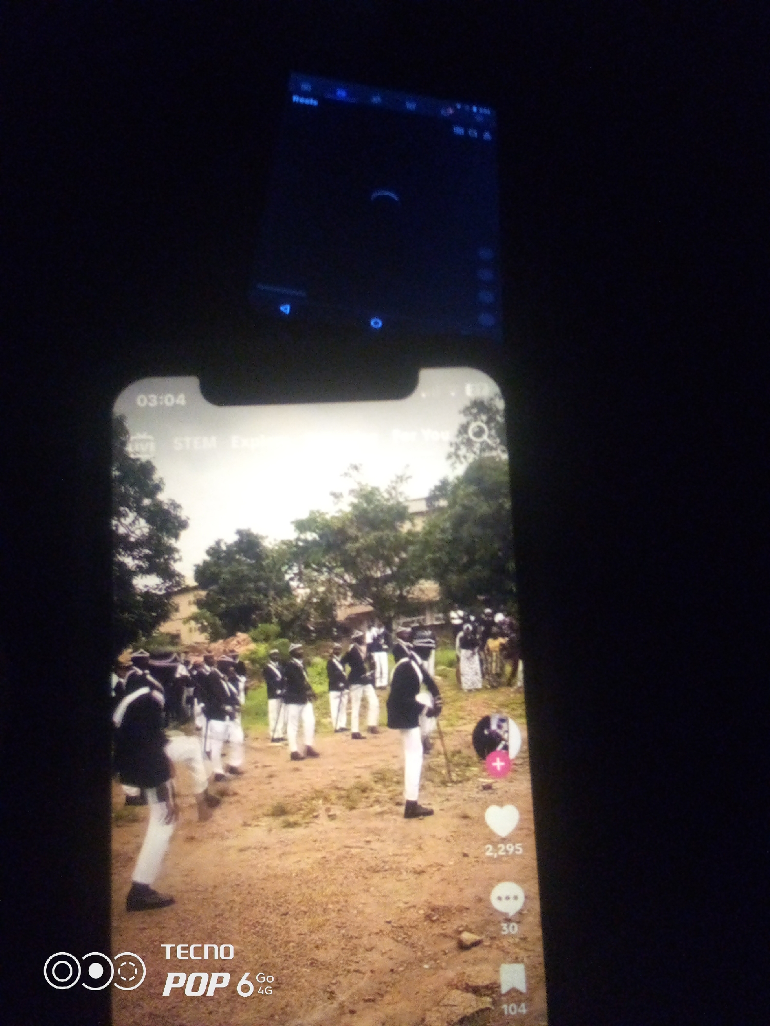 The image displays a person holding a smartphone which is capturing a video. The video on the smartphone's screen shows a group of individuals in uniform, appearing to be in a parade or ceremony, in an outdoor setting with trees and buildings in the background. The scene is dimly lit. The lower portion of the smartphone screen shows social media interface elements, including like and comment counts. The top part of the image is dark, showing the screen of another device or the reflection of the surrounding environment. There are no visible indicators in the image that place the scene specifically in Howayi, Nigeria.