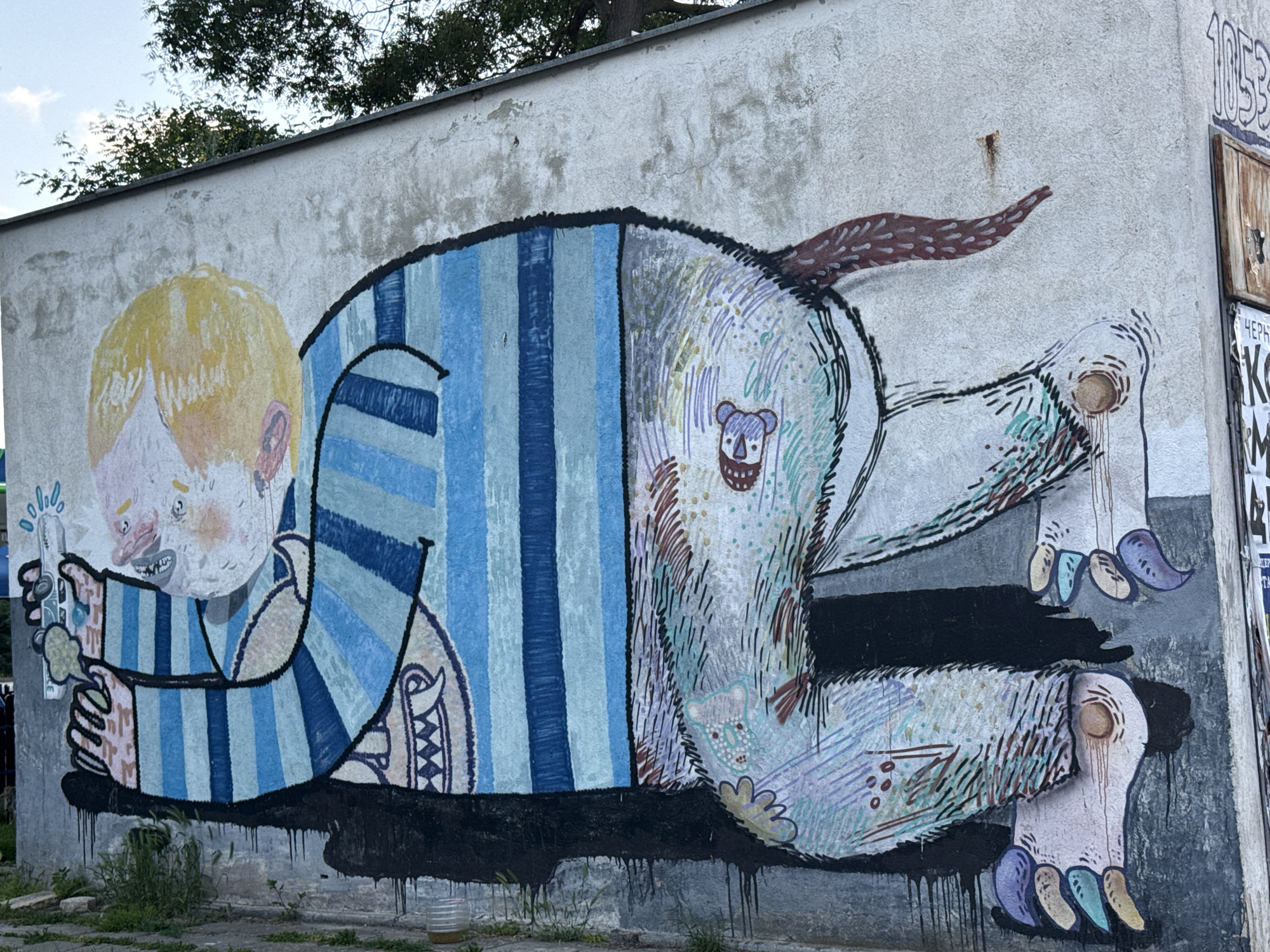 The image shows a mural on a concrete wall located in Unknown, Unknown. The mural depicts a stylized figure that appears to be a combination of a child and a bear. The figure's head is that of a child with blonde hair and exaggerated facial features, holding a small object in its hand. Its body is large and striped in shades of blue, resembling a prisoner's uniform. The rear portion of the figure transitions into the fur and paws of a bear, complete with claws and a tail. A small bear face is depicted on the back. The mural is painted with thick lines and a muted color palette. The wall shows signs of age and weathering, with cracks and discoloration. Black paint drips down from the bottom edge of the mural onto the ground. Patches of grass and weeds grow near the base of the wall.