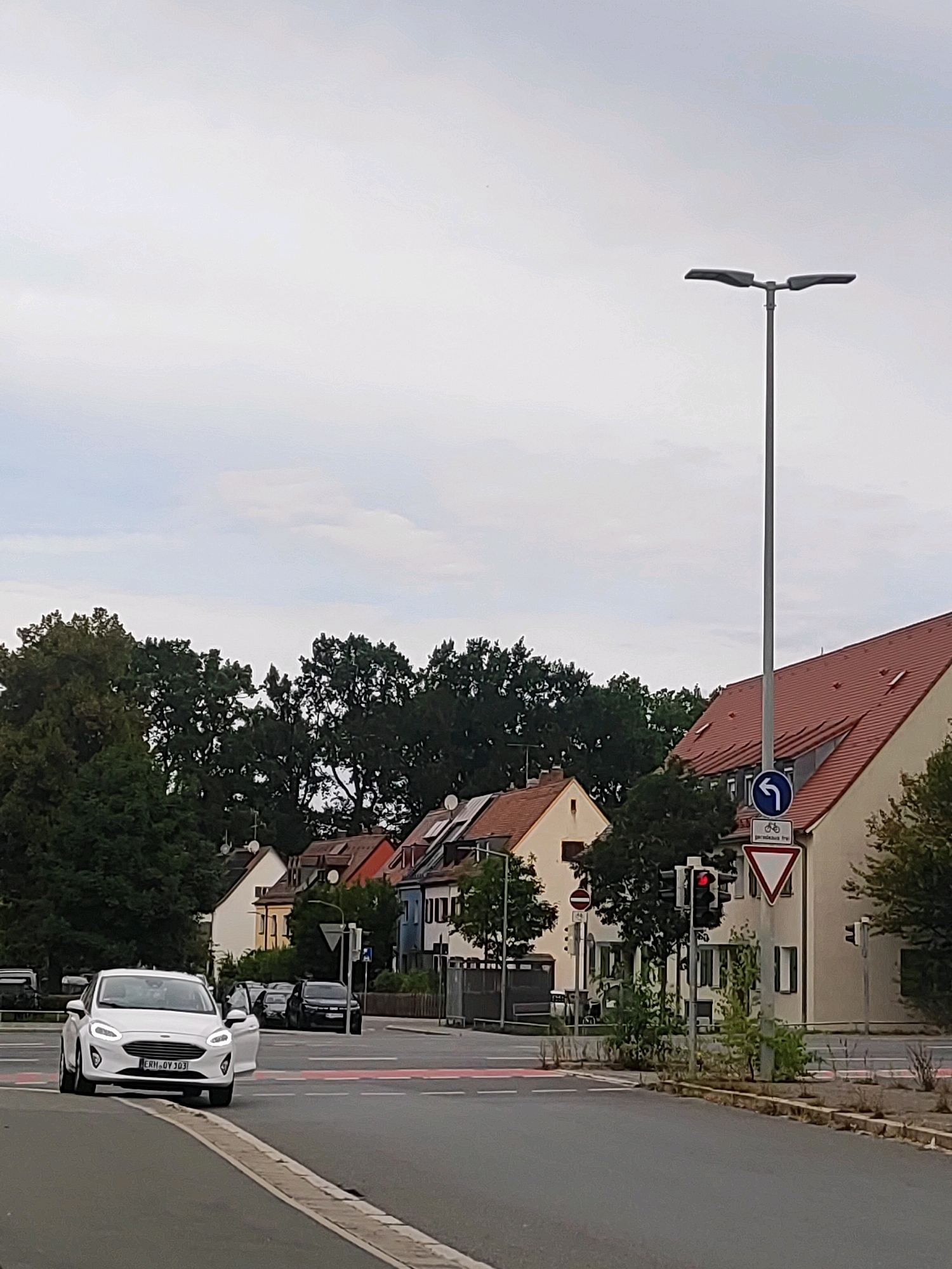 The image captures a street scene in Nuremberg, Germany, with a white Ford Fiesta car in the foreground. The car, with license plate ERH-QY-103, is turning left at an intersection. The intersection features a pedestrian crossing and traffic lights, with the red light illuminated. Behind the car, a dark-colored SUV is visible, also at the intersection. In the background, a line of residential buildings with red-tiled roofs stretches along the street, interspersed with mature trees. A tall, modern street lamp with two lights stands prominently on the right side of the road, near the buildings. Traffic signs, including a blue circular sign with a white arrow indicating a left turn and a symbol for bicycles, as well as a yield sign, are present near the intersection. The sky is overcast, suggesting a cloudy day. The overall environment appears to be a suburban or residential area of Nuremberg.