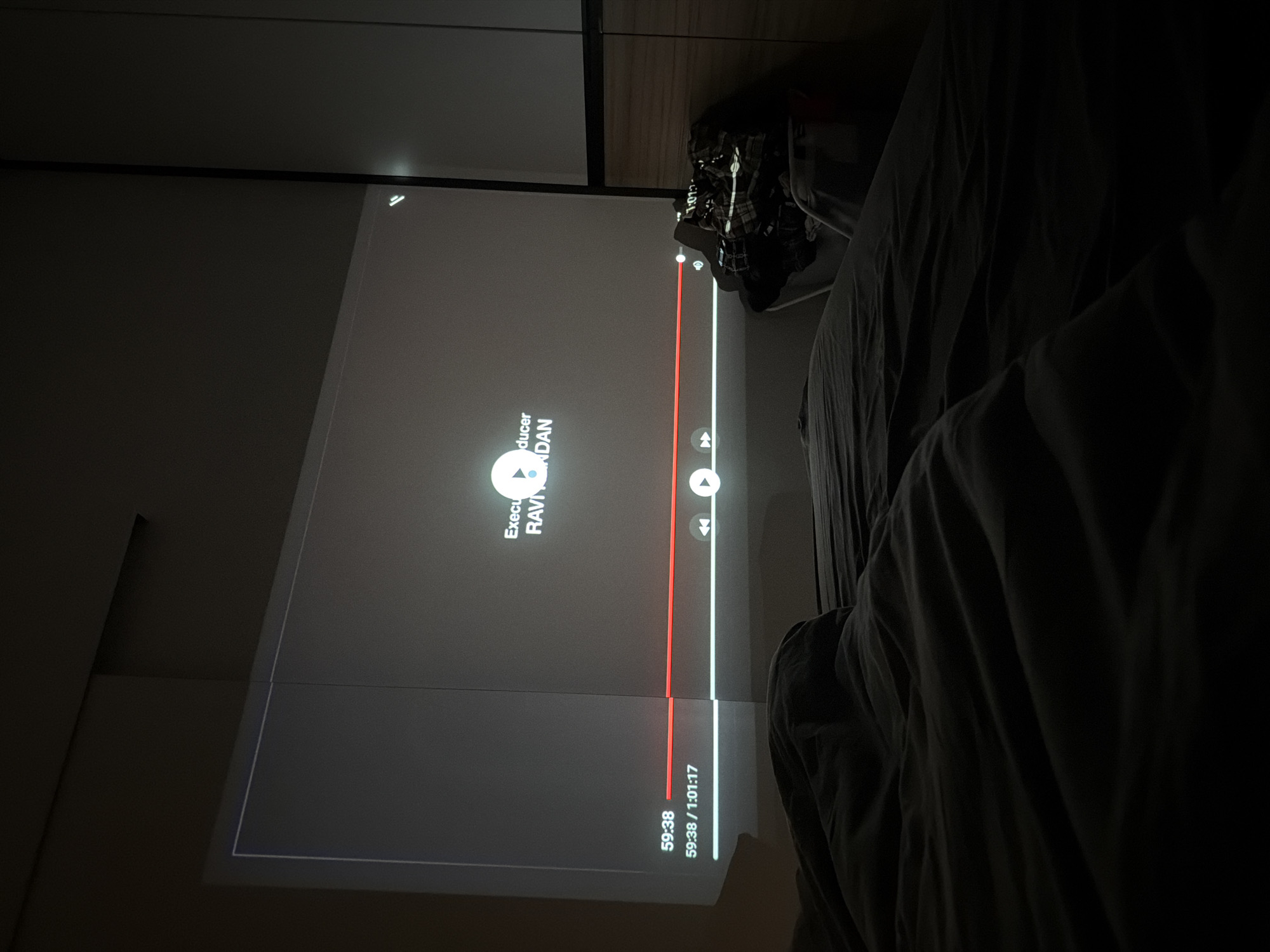 The image shows a dark room with a screen displaying a paused video. The video title is not fully visible, but the visible part reads "Execu...ducer RAVI...NDAN". The video is at the timestamp 59:38 out of 1:01:17. To the right of the screen, a bed with dark grey linens is visible. The room appears to be dimly lit or dark, with the screen providing the primary source of light. The location is not evident from the image, and cannot be determined based on available details.