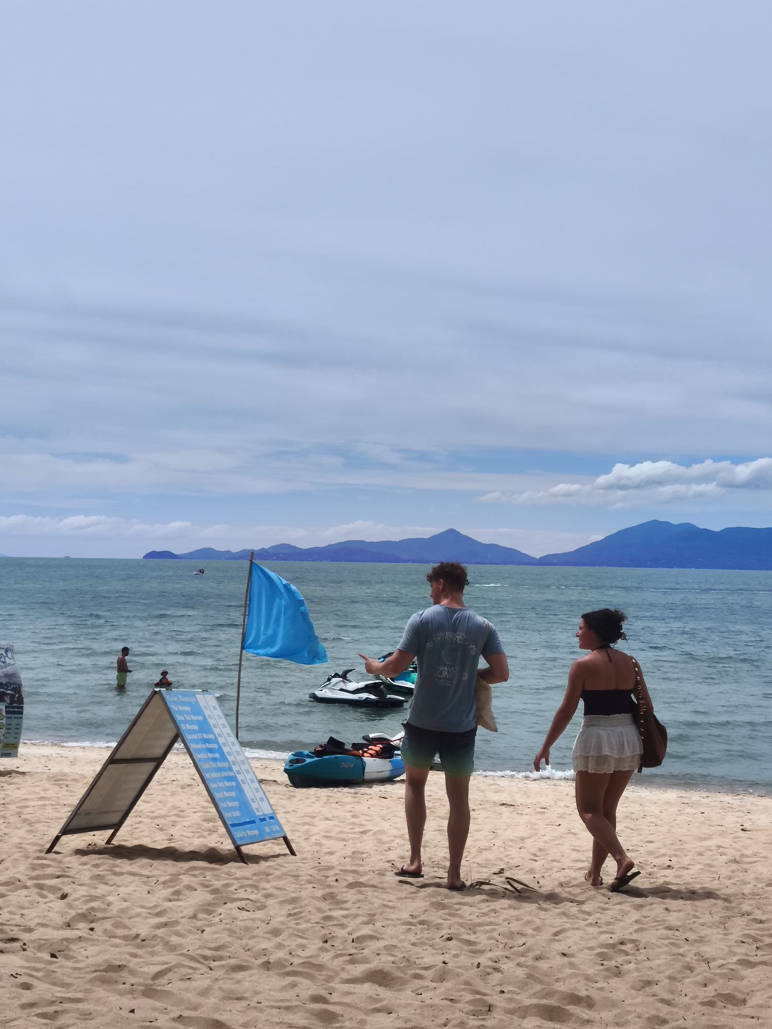 A man and a woman stand on a sandy beach in Baan Mae Nam, Thailand, facing away from the camera. The man wears a blue t-shirt and patterned shorts, while the woman wears a black strapless top and a white skirt. They appear to be walking towards the ocean.
In the background, the blue ocean stretches towards a hazy coastline with mountains. Several jet skis are visible near the shore, and a blue flag is planted in the sand. A triangular sign board displaying various massage services and prices is positioned in the foreground. Two people are wading in the water in the mid-ground, and a kayak with life vests is near the shore. The sky is overcast with scattered clouds.