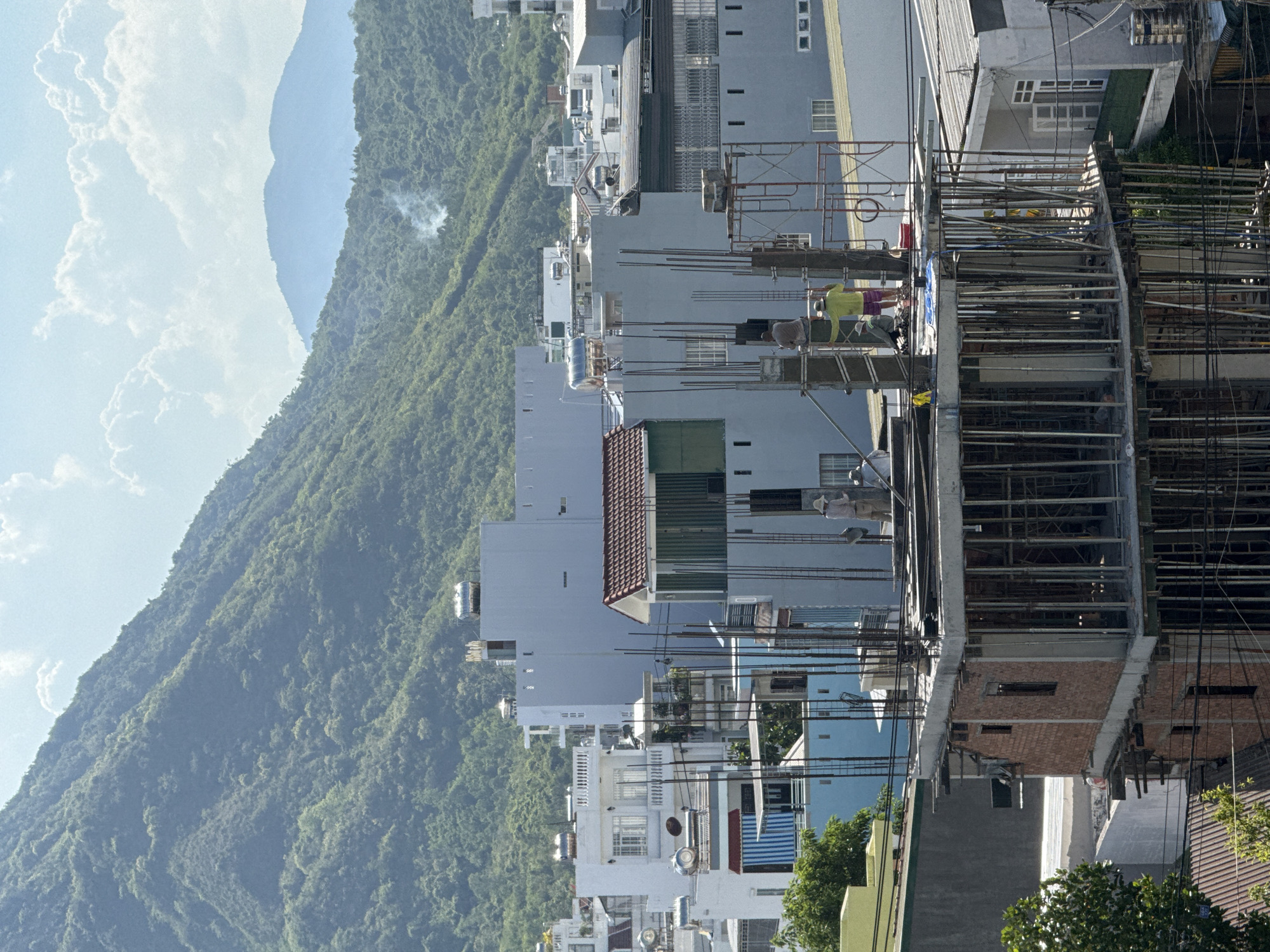 The image depicts an urban construction site in Bac Nha Trang Ward, Vietnam, set against a backdrop of lush green mountains. In the foreground, a multi-story building is under construction, characterized by exposed brickwork, a concrete frame, and numerous vertical steel reinforcing bars. Scaffolding surrounds parts of the structure. Two construction workers are visible on the upper levels of the partially built edifice, one wearing a yellow shirt and the other in a beige hat and shirt.
Behind the construction site, a dense cluster of buildings, primarily residential, stretches across the landscape. Many of these buildings are white or light-colored and feature various architectural styles, including balconies and what appear to be solar water heaters on their rooftops. The middle ground features more substantial, light blue and grey buildings, some with visible windows and balconies. A section of a building with a red-tiled roof and green siding is also present.
The background is dominated by steep, verdant mountains covered in dense vegetation, rising towards a bright blue sky with scattered white clouds. In the distance, faint mountain ranges are visible, and a wisp of smoke rises from the mountainside, suggesting distant activity. The overall scene conveys a sense of ongoing development within a naturally beautiful, hilly environment.