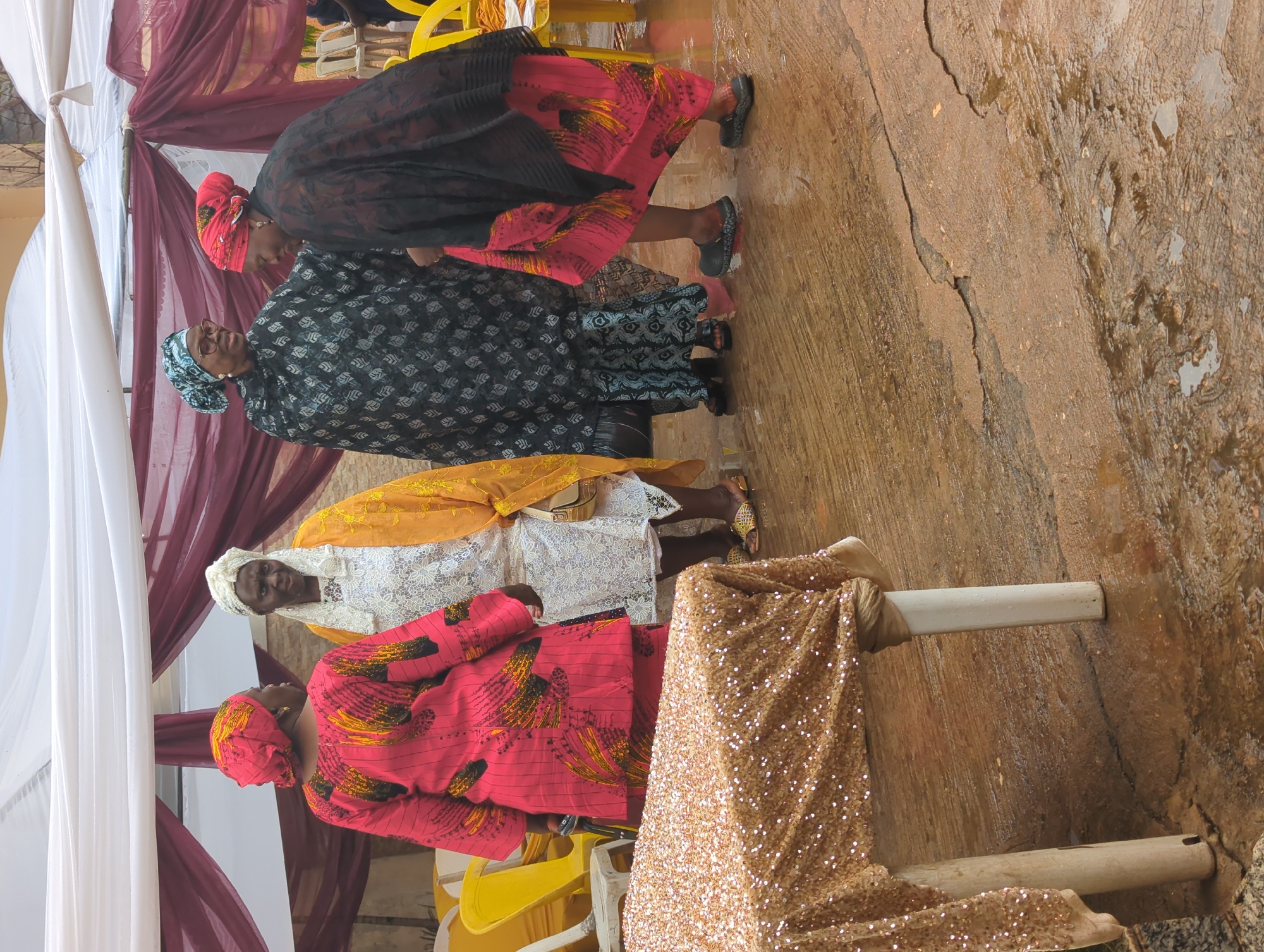 The vertically oriented image depicts five women in colorful traditional attire gathered under a canopy, likely at an outdoor event in Unguwar Rimi, Nigeria.
In the foreground, a woman wears a vibrant red gown with yellow and black patterns, complemented by a matching head wrap. To her left, another woman is dressed in a white lace top over a yellow garment, with a yellow head wrap, and offers a slight smile towards the viewer. Behind them, a third woman wears a dark patterned dress and a blue and green patterned head wrap, along with spectacles, seemingly engaged in conversation or observation. Further back, two more women are visible: one in a black outfit with a red and yellow patterned skirt and a red head wrap, and another in a dark patterned dress with a patterned head wrap. All women are wearing flat shoes or sandals.
The setting is an informal outdoor space, possibly a courtyard, with a canopy constructed from white and maroon fabrics providing shade. The ground is a prominent detail, appearing as rough, wet earthen ground with visible cracks and puddles, indicating recent rain or damp conditions. To the right, a shimmering gold sequined fabric is draped over white poles, serving as decorative elements for the event. Yellow plastic chairs are partially visible in the background, suggesting a seating arrangement for attendees. No discernible text is present in the image. The scene conveys a sense of communal gathering or a social occasion taking place during the daytime.
