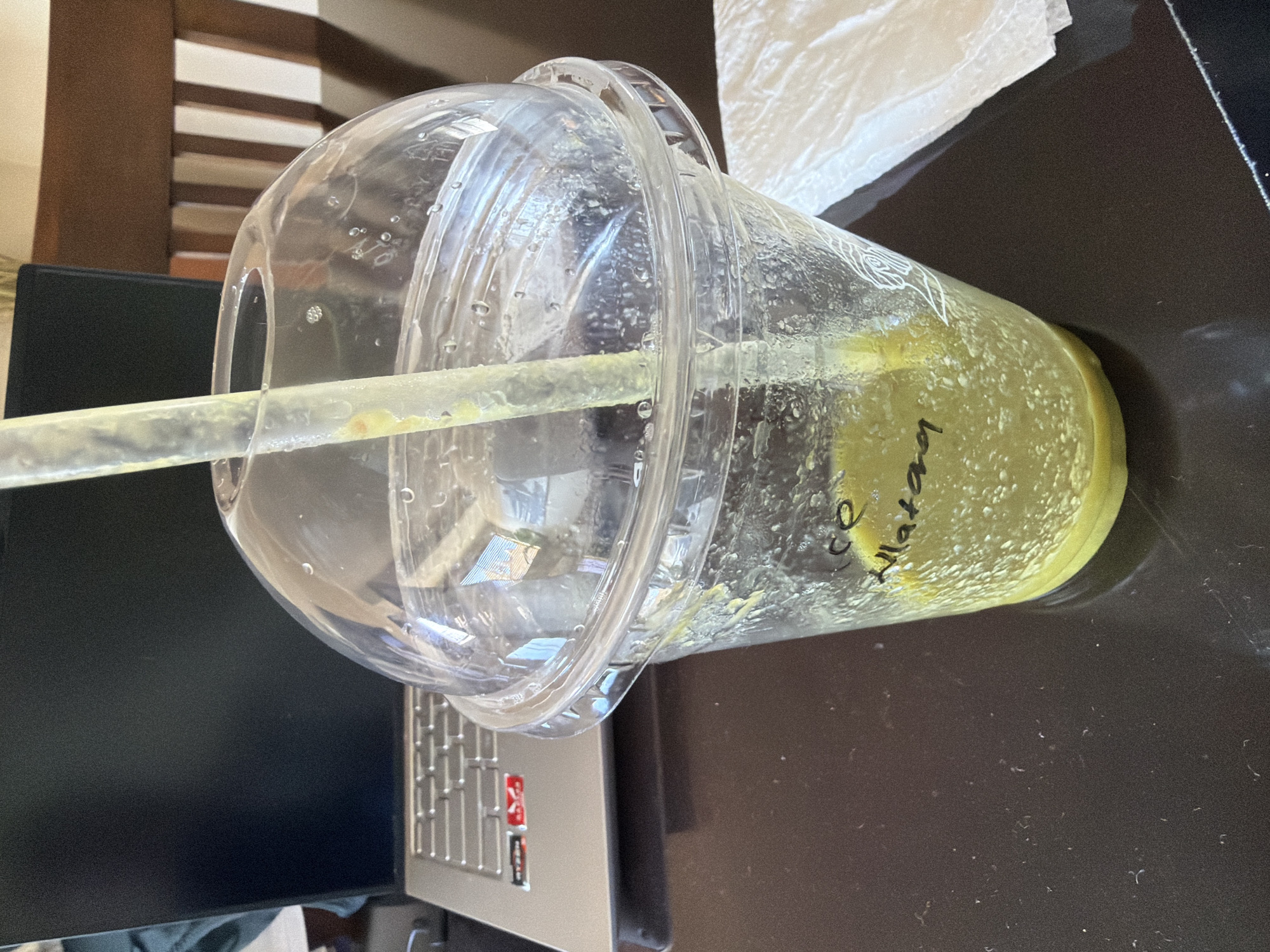 A clear plastic cup with a dome lid and a straw sits on a dark brown surface, likely a desk or table. Inside the cup, a greenish-yellow beverage is visible, possibly a smoothie or juice, with handwritten text in black marker on the side of the cup near the liquid level. The text appears to read "CE Matan". To the left of the cup, a laptop or monitor screen is partially visible, with a portion of the laptop's keyboard and chassis also in view. A white paper napkin is positioned behind the cup. In the background, a wooden cabinet or shelving unit with vertical stripes is seen. The specific location of Cape Town, South Africa cannot be inferred from the image.
