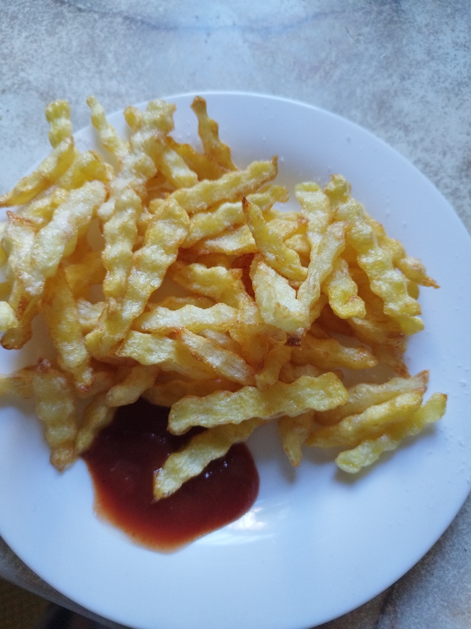 A white plate with a pile of golden-brown crinkle-cut french fries and a dollop of dark red ketchup is placed on a textured, light-colored surface. The french fries are seasoned with salt and appear crispy. The background is out of focus, but suggests a casual dining setting. The image does not provide any information about the location of Lozova, Ukraine.