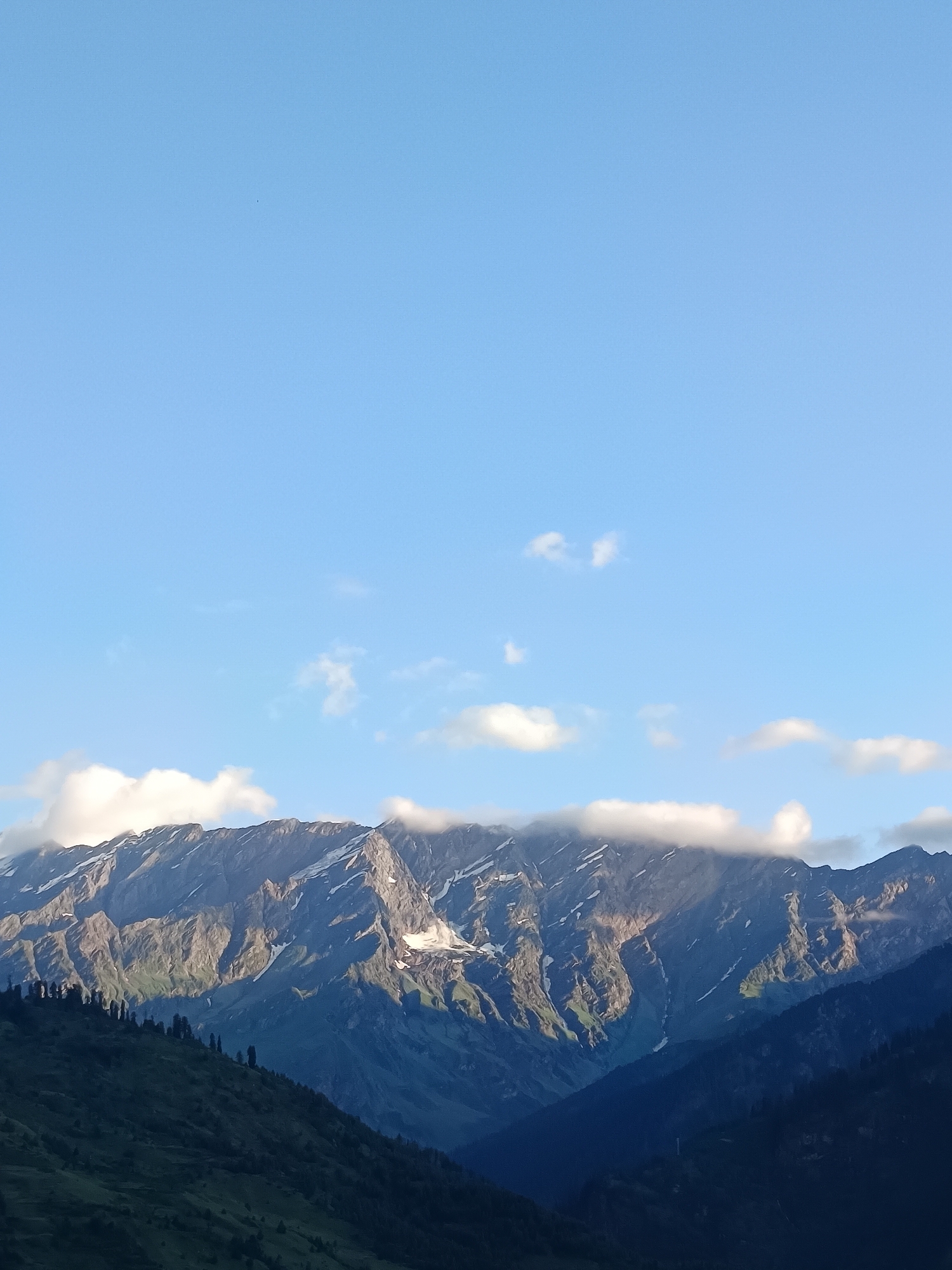 The image captures a panoramic view of majestic, snow-capped mountains under a clear blue sky with scattered white clouds. The mountain range dominates the lower half of the frame, showcasing rugged peaks and deep valleys. Patches of snow are visible on the higher elevations, contrasting with the dark, rocky slopes and lush green vegetation in the foreground. The lighting suggests it is either early morning or late afternoon, casting shadows that accentuate the mountainous terrain. In the distance, a few wispy clouds drift across the sky, adding depth to the scene. The overall impression is one of natural grandeur and tranquility. The specific location is Vashisht, India, a region known for its picturesque Himalayan landscapes.