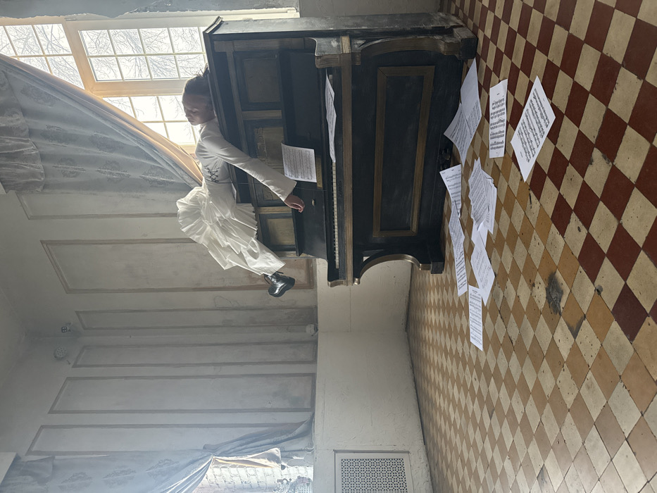 This image depicts an interior scene within Moscow, Russia, characterized by classical architecture. A single individual is positioned horizontally across the top of an antique, dark-toned upright piano with gold/brass accents. The person, dressed in a long-sleeved white top and voluminous white ruffled trousers, lies face down or sideways, with their head resting on the piano's top surface. Their left leg is bent at the knee, wearing a black chunky-soled boot, while their right arm hangs loosely down towards the piano keys. One sheet of music rests on the piano's music stand. Numerous sheets of printed music are scattered across the checkered floor in front of the piano.
The room features light-colored walls adorned with ornate classical molding panels. The floor is composed of a checkerboard pattern of aged light brown/beige and reddish-brown square tiles, showing signs of wear and staining. Two large windows dominate the background; the right window, partially obscured by light blue patterned drapes, reveals bare tree branches outside. The left window, also draped, shows a brick exterior and features a white decorative vase or statuette on its sill. A white patterned grate is visible on the lower left wall, and a small, white, round device is mounted high in the corner of the room. The scene is illuminated by natural light entering through the windows.