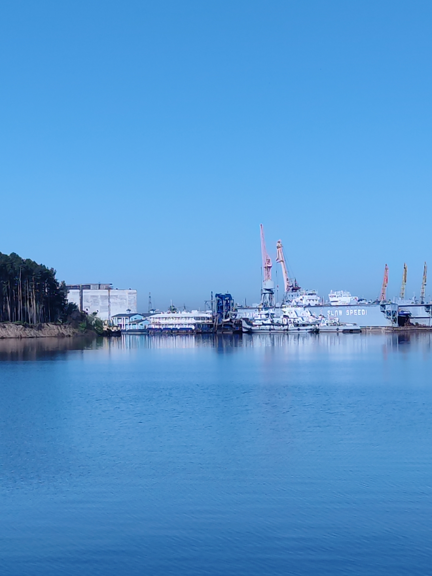 The image shows a waterfront scene in an unknown city. A body of water dominates the foreground, reflecting the clear blue sky. In the middle ground, there are several buildings and industrial structures lining the shore. Cranes of varying colors are visible, suggesting a port or shipbuilding area. Multiple boats and ships are docked along the water. A line of trees runs along the edge on the left side, next to a large building. The overall scene suggests a working port with industrial and commercial activity.