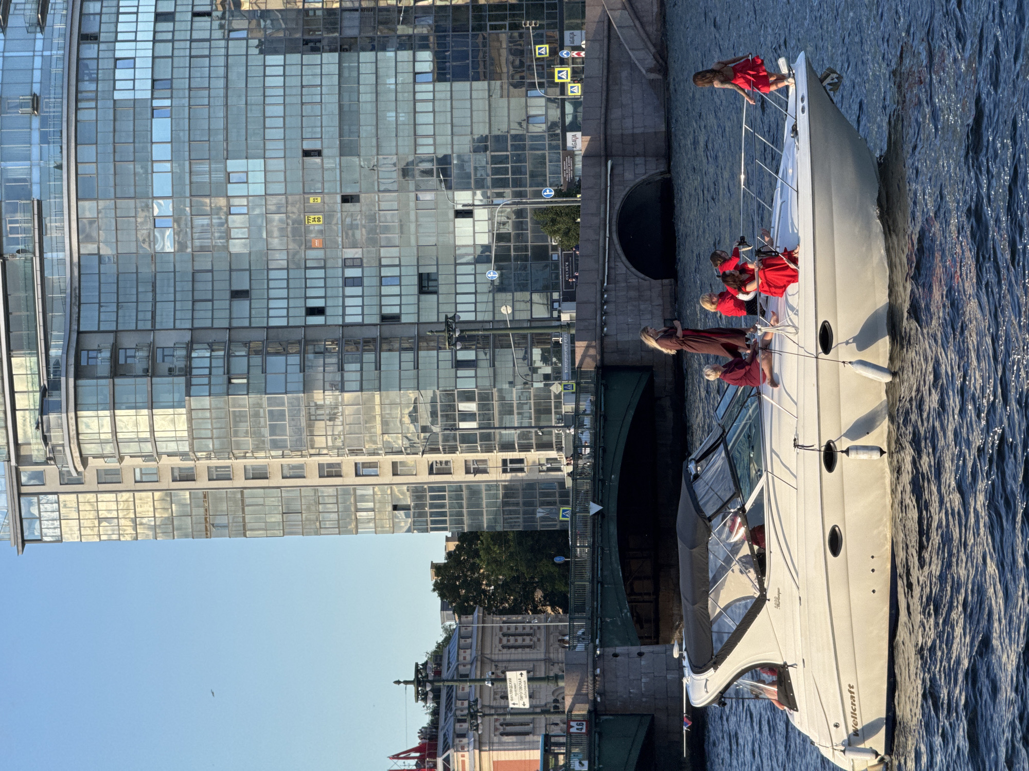A white motorboat with a black canopy travels on a body of water, likely a river, in Saint Petersburg, Russia. Six people are visible on the boat, dressed in red and maroon attire. The boat has the name "Wellcraft" visible on its side. In the background, a modern glass-covered building with multiple floors and balconies reflects the sky. A green bridge with stone arches spans the water, and further in the background, older stone buildings and a lamppost are visible. The sky is clear and blue, indicating a sunny day. The scene appears to be a leisurely boat ride in an urban setting.