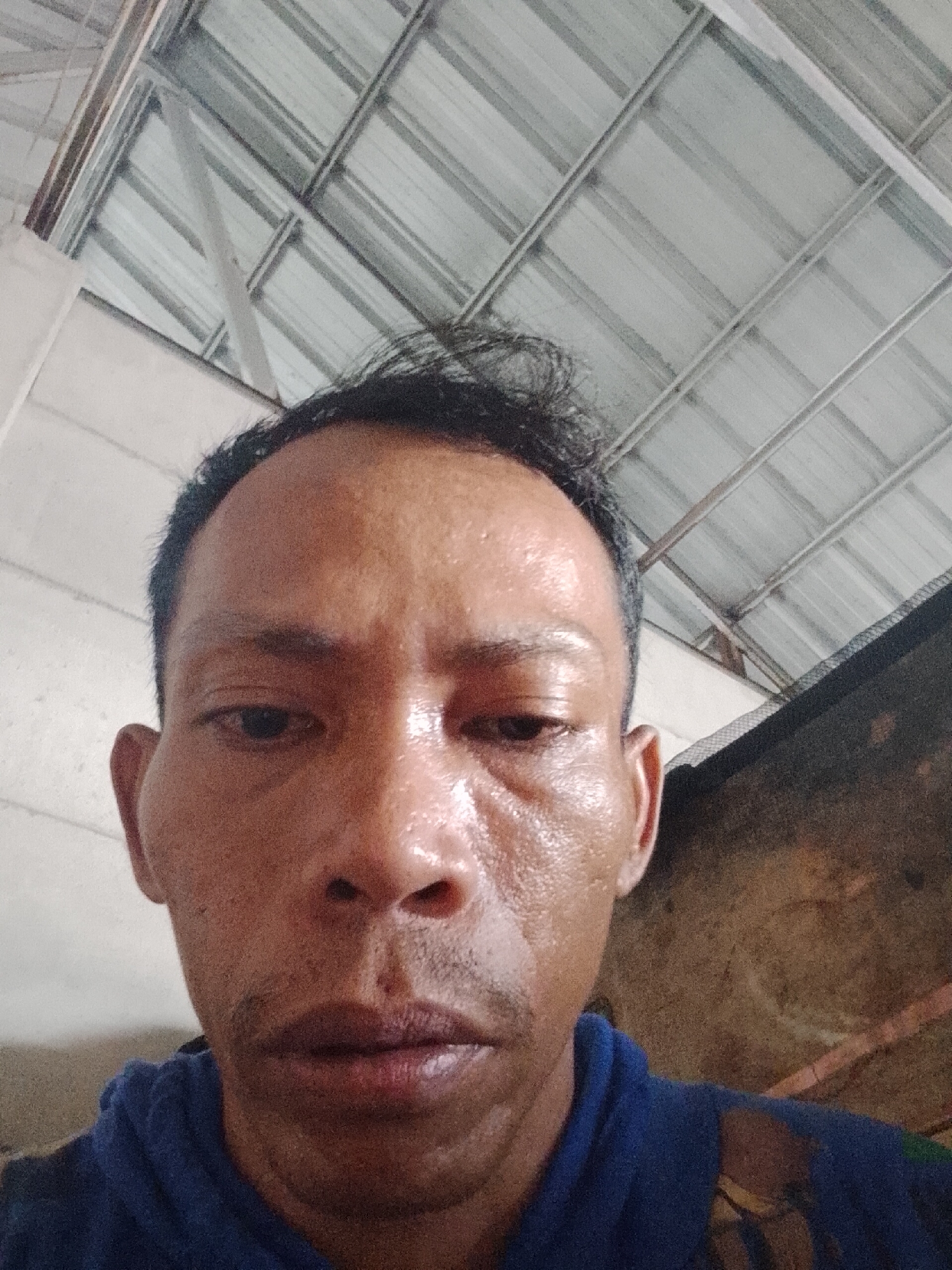 A close-up, low-angle portrait of a middle-aged Asian man with short black hair, facing the camera. His skin appears damp or sweaty. He has dark eyes, a wide nose, and a thin mustache. He is wearing a blue garment. The background shows a metal roof structure, possibly of a simple building or shelter. The setting is potentially the city of Bukit Ketok, Indonesia.