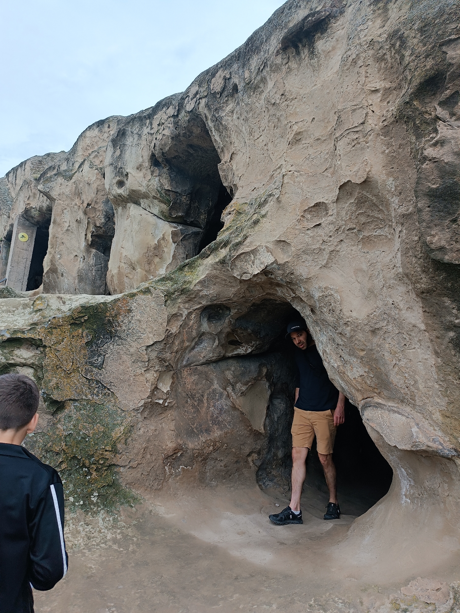 The image depicts two individuals exploring the cave city of Uplistsikhe, Georgia. In the foreground, a person with short brown hair, wearing a black jacket with white stripes on the sleeves, is viewed from the back, facing the caves. To the right, a man wearing a black t-shirt, a black baseball cap, and tan shorts stands partially inside a cave opening, looking towards the camera. The environment is characterized by large rock formations with natural cave entrances, suggesting an ancient historical site. The ground is sandy and uneven. The sky is overcast and light in color.