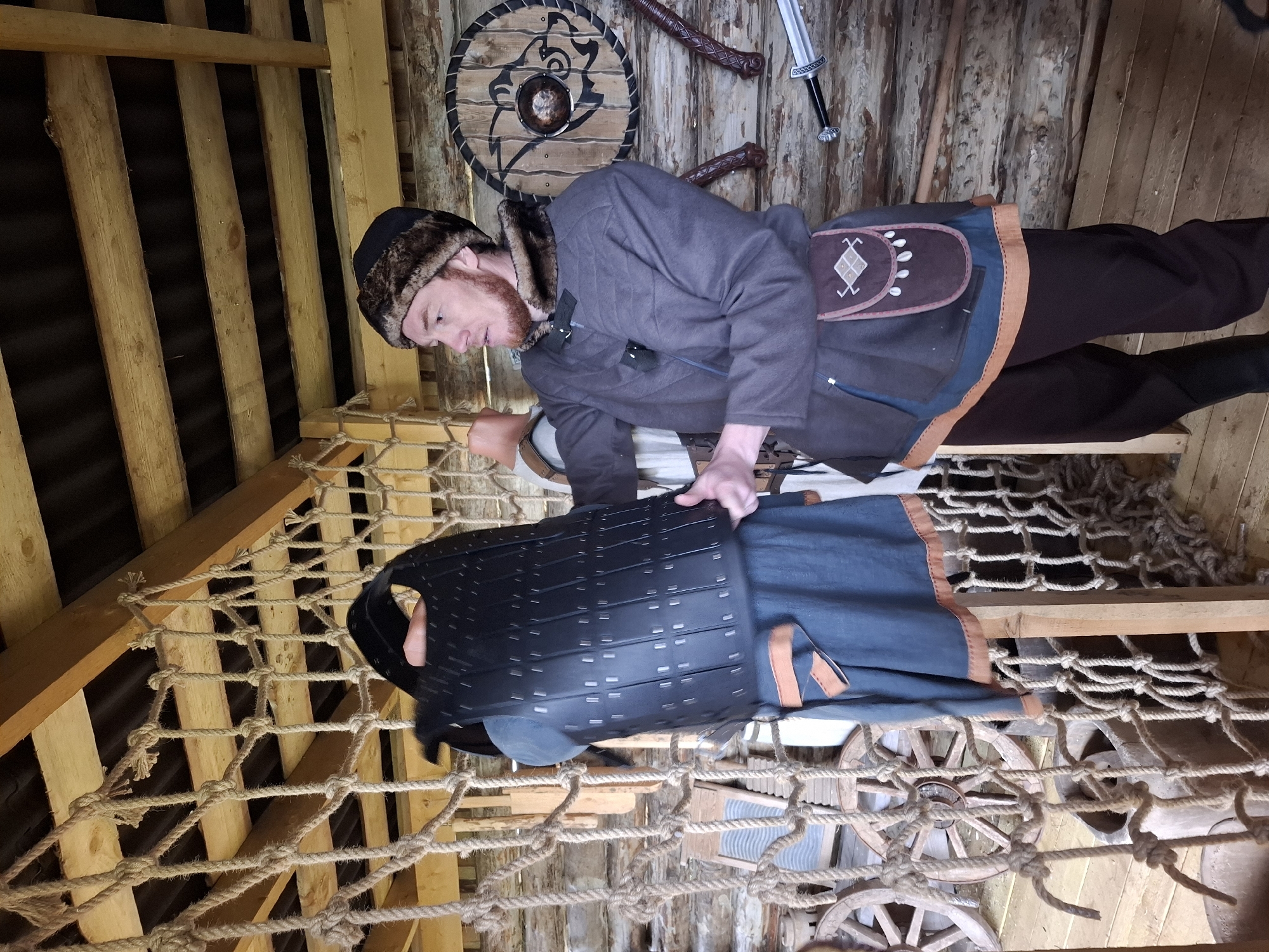 A medium shot captures a man indoors, likely in a reconstructed medieval or Viking-era setting. He is wearing a dark gray coat over a brown tunic and pants, and a fur-trimmed hat. A pouch with a distinctive design is strapped to his side. He is adjusting what appears to be a piece of dark, possibly leather or composite armor, laid out over a wooden net strung across timber supports. The background includes a wall constructed of rough-hewn logs and planks, adorned with a shield, weapons, and other objects evocative of the period. The location is the city of Адам, Russia. The overall scene suggests a historical reenactment, museum display, or thematic restaurant or entertainment venue.