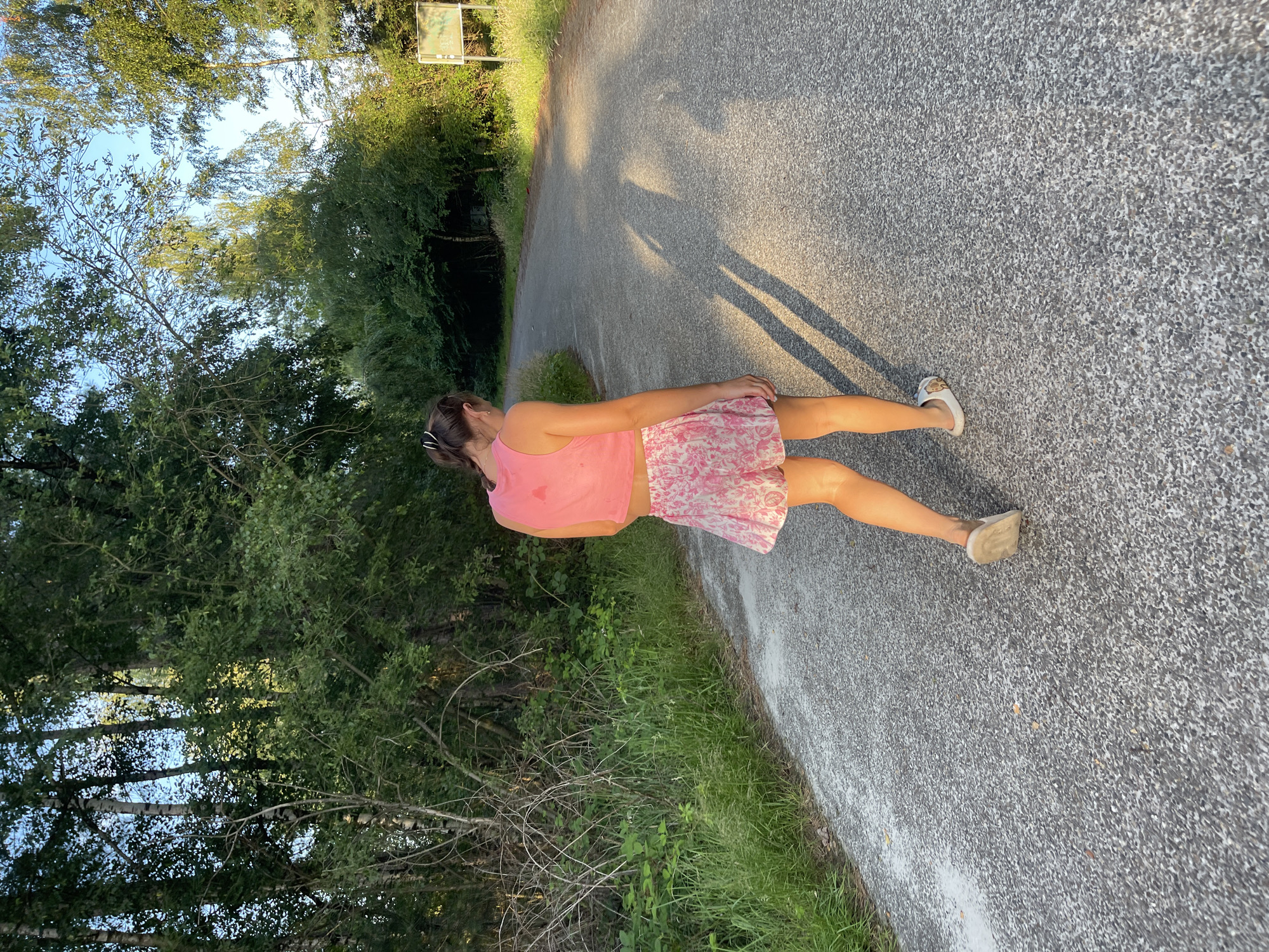 A young person with brown hair pulled back in a ponytail, wearing a pink tank top and a floral print skirt, walks away from the camera on a paved road. The person is wearing white slip-on sandals. The road curves to the right and is surrounded by dense green trees and vegetation. The sun is low in the sky, casting long shadows on the road. There is a signpost visible on the right side of the road in the distance. The environment appears to be a rural or park-like setting.