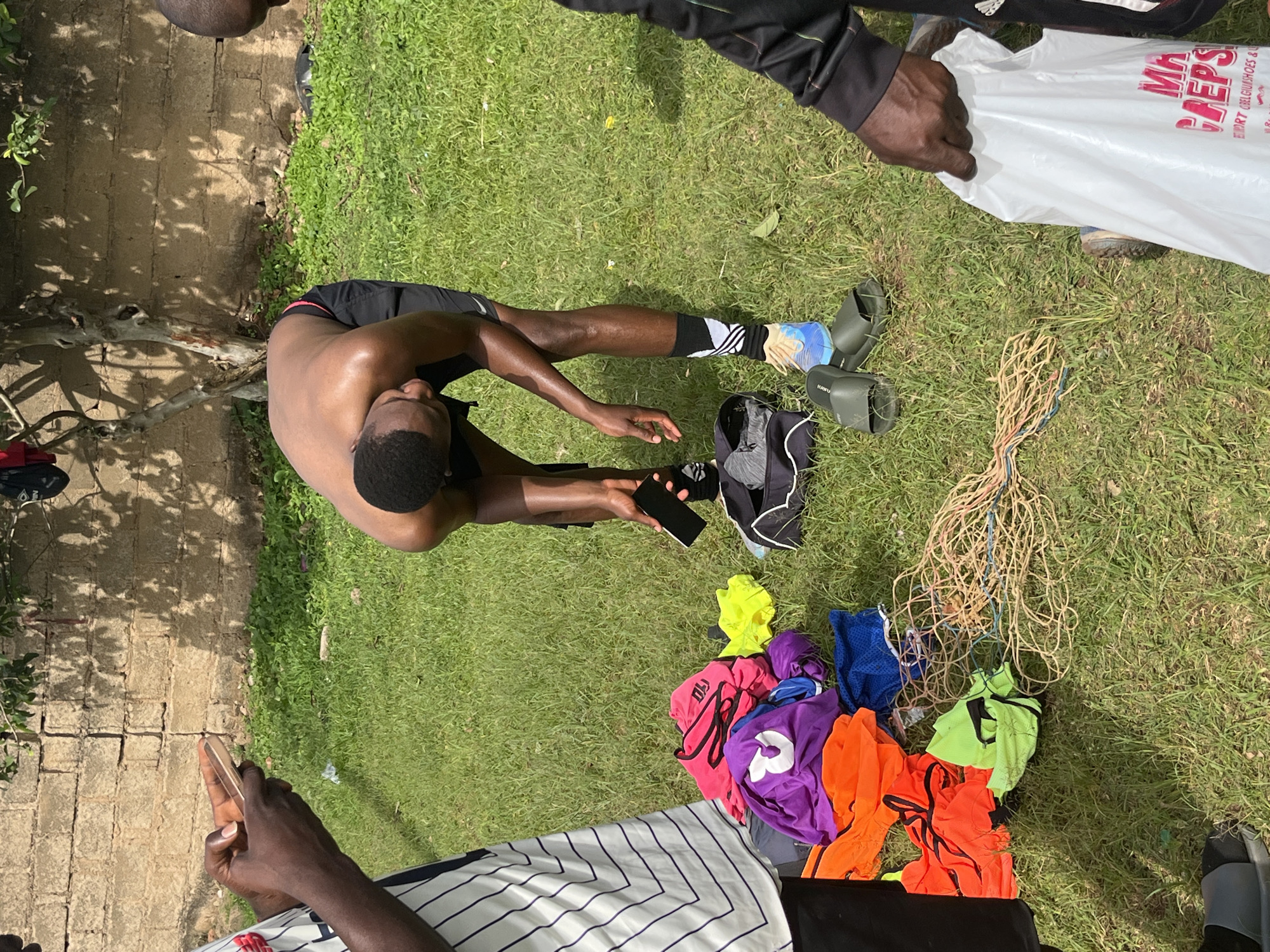The image depicts an outdoor scene on a grassy area with a brick wall in the background. The location is identified as Jos, Nigeria. Several people are present, though not all are fully visible. One individual, a shirtless young Black man, is bent over, holding a black smartphone. Around him, various items are scattered on the grass, including colorful sports bibs (purple, orange, pink, green, yellow, blue), a black duffel bag, a tangled rope, and a pair of dark green slides. Another person on the left is partially visible, holding a gold-colored smartphone and wearing a white and black striped shirt. To the right, another individual is holding a white plastic shopping bag with red lettering. The overall atmosphere suggests a casual gathering or a break during an outdoor activity.