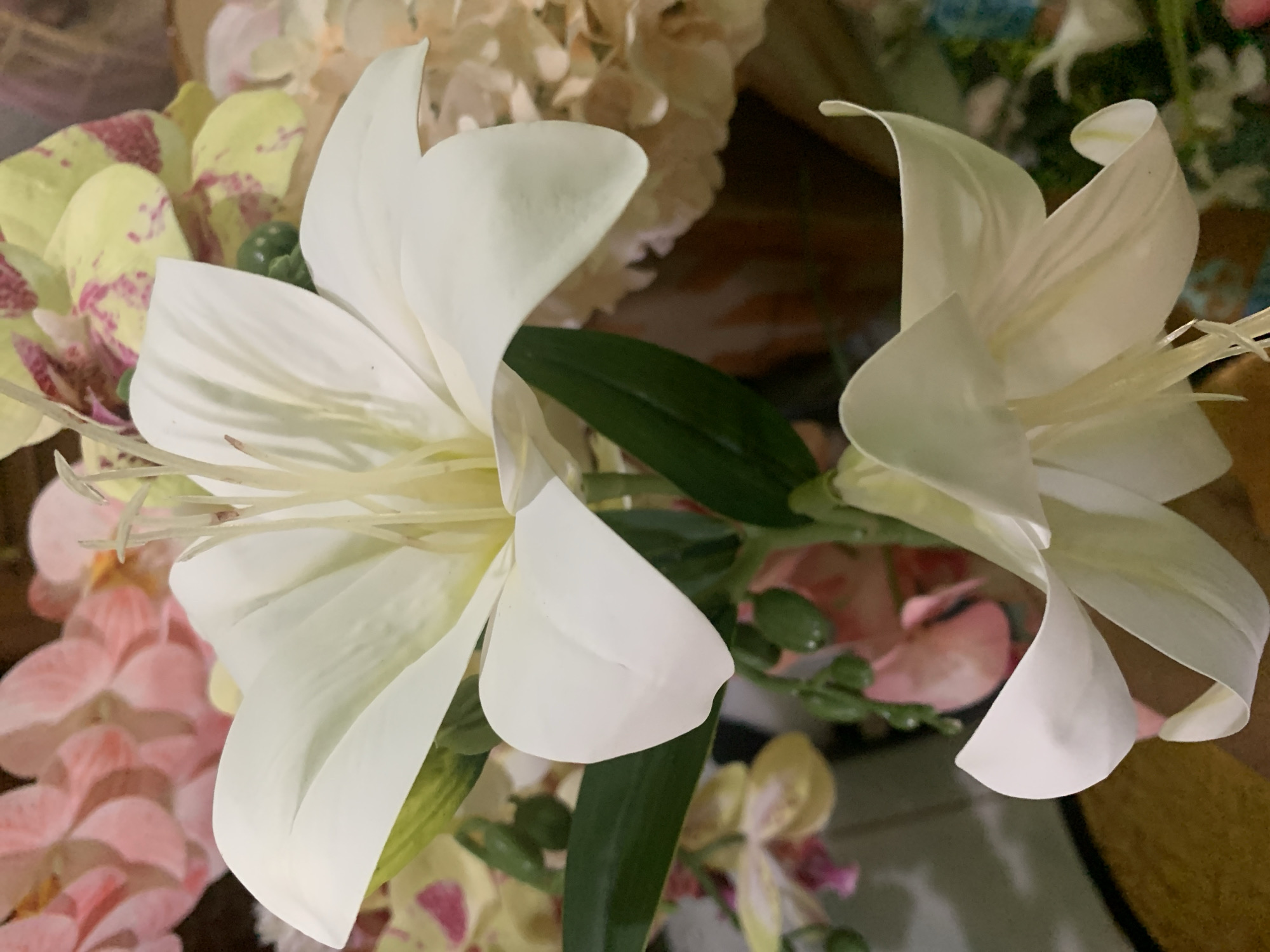 A close-up, shallow depth-of-field shot focuses on two artificial white lilies with pale yellow stamens. The larger lily is in the upper center of the frame, its petals unfurled. Below and to the right, a second, slightly smaller lily is also visible. The background is a blur of soft pink, pale yellow with pink speckles, and muted peachy tones, suggesting other artificial flowers. Green leaves and buds are scattered among the lilies. The lighting is soft and diffuse, creating a gentle illumination. The image does not depict any identifiable landmarks or environmental features that would allow for its localization to Kuala Pembuang, Indonesia, or any other specific city.