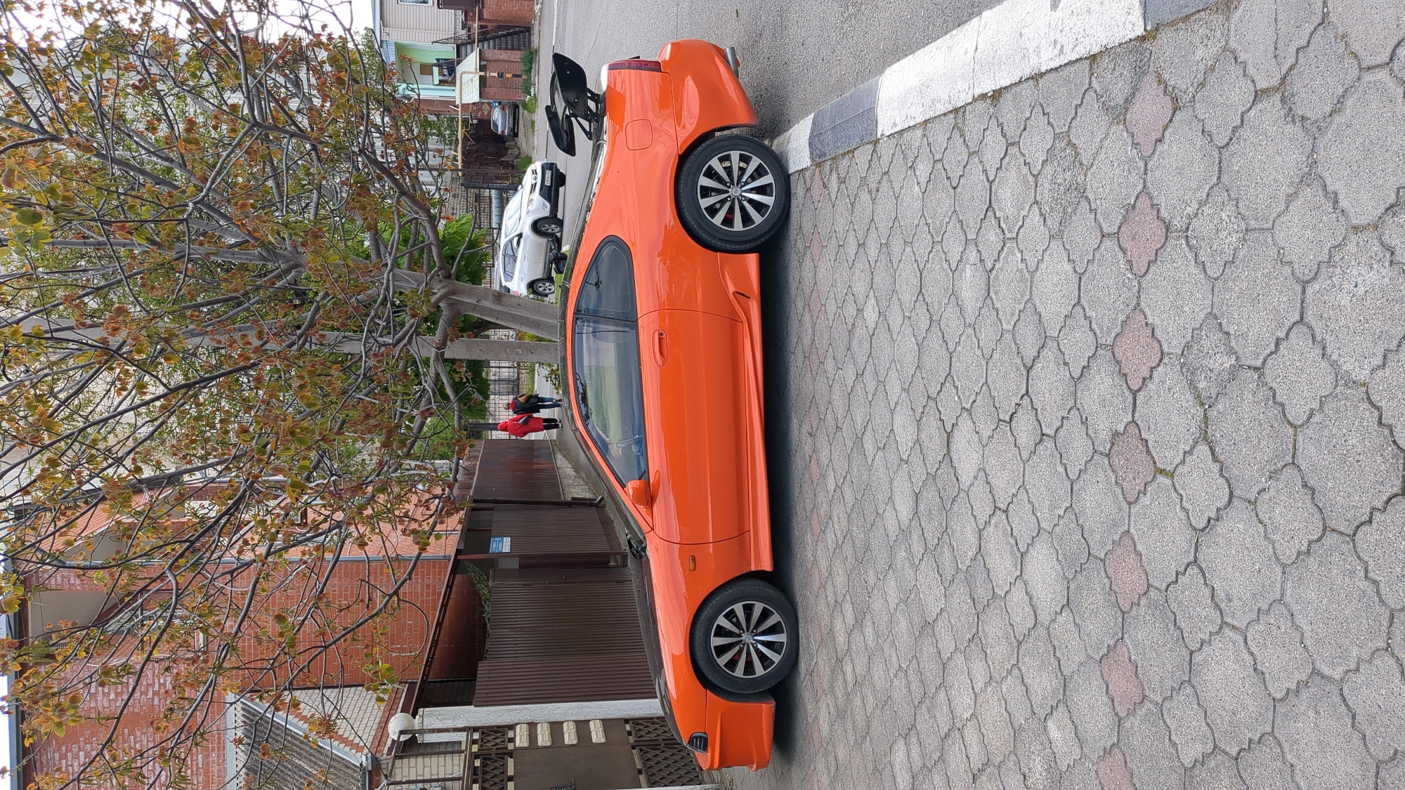 The image shows an orange coupe car parked on a cobblestone street with a white curb. The car has dark-tinted windows and aftermarket wheels. It is near a tree with autumn leaves, a brown fence, and brick buildings. In the background, other cars are parked on the street. The location is Gelendzhik, Russia. The image includes elements suggesting an urban residential area.