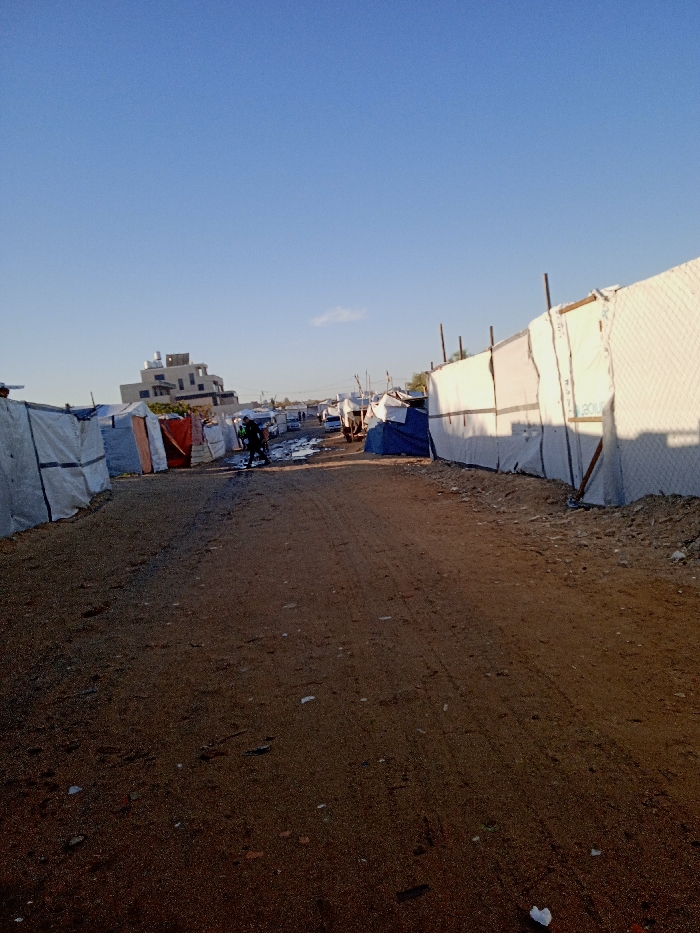 This image depicts an unpaved dirt path through a displacement camp in Khan Yunis, Palestinian Territory, under a clear blue sky with a single distant cloud. On the left, a row of makeshift shelters is constructed from various materials including tarpaulins and plastic sheeting. On the right, a long barrier or fence line is composed of white sheeting, some displaying the "UNICEF" logo, interspersed with chain-link fencing, all supported by vertical wooden poles. Shadows are cast towards the left side of the path by the right-side barrier. In the mid-ground, a solitary individual in dark clothing, possibly a dark green or black jacket, walks away from the camera along the path. Further down the path, the ground appears muddy and wet, and additional shelters are visible alongside what appear to be several white vehicles, possibly vans or trucks. In the far background, a multi-story concrete building with a rooftop water tank stands prominently above the temporary structures. Debris, including a small white object, is scattered on the dusty foreground.