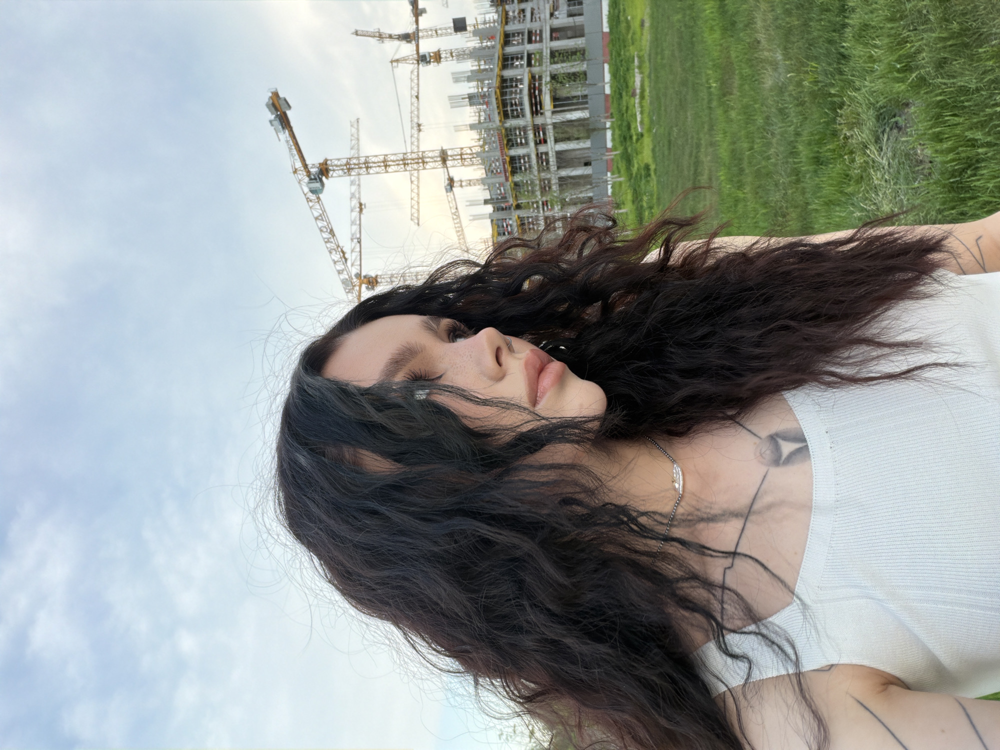 A low-angle shot depicts a woman with long, dark, curly hair and freckles. She wears a white top and a silver necklace. Visible tattoos adorn her chest and arm. The backdrop features a construction site with several yellow cranes and a tall building structure, possibly in Moscow, Russia. The sky is overcast, and a patch of green grass is also visible. The image is slightly rotated.