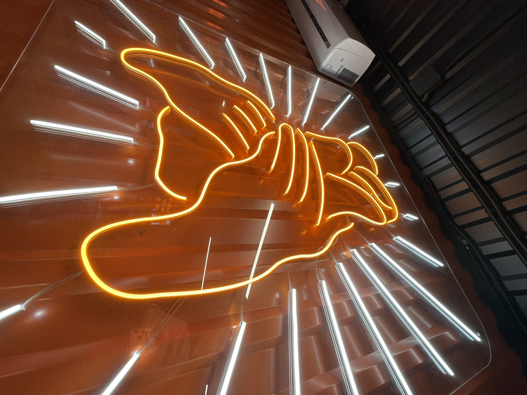 A low-angle shot captures a neon sign attached to a wall. The sign features a stylized depiction of hands, illuminated with a bright orange-yellow neon light. White neon "rays" emanate from the hands, creating a starburst effect. The sign is mounted on an orange-brown wall with horizontal ribbing. The ceiling is dark with metal grating. An air conditioning unit is mounted near the top of the image. The location is Krasnoyarsk, Russia.
