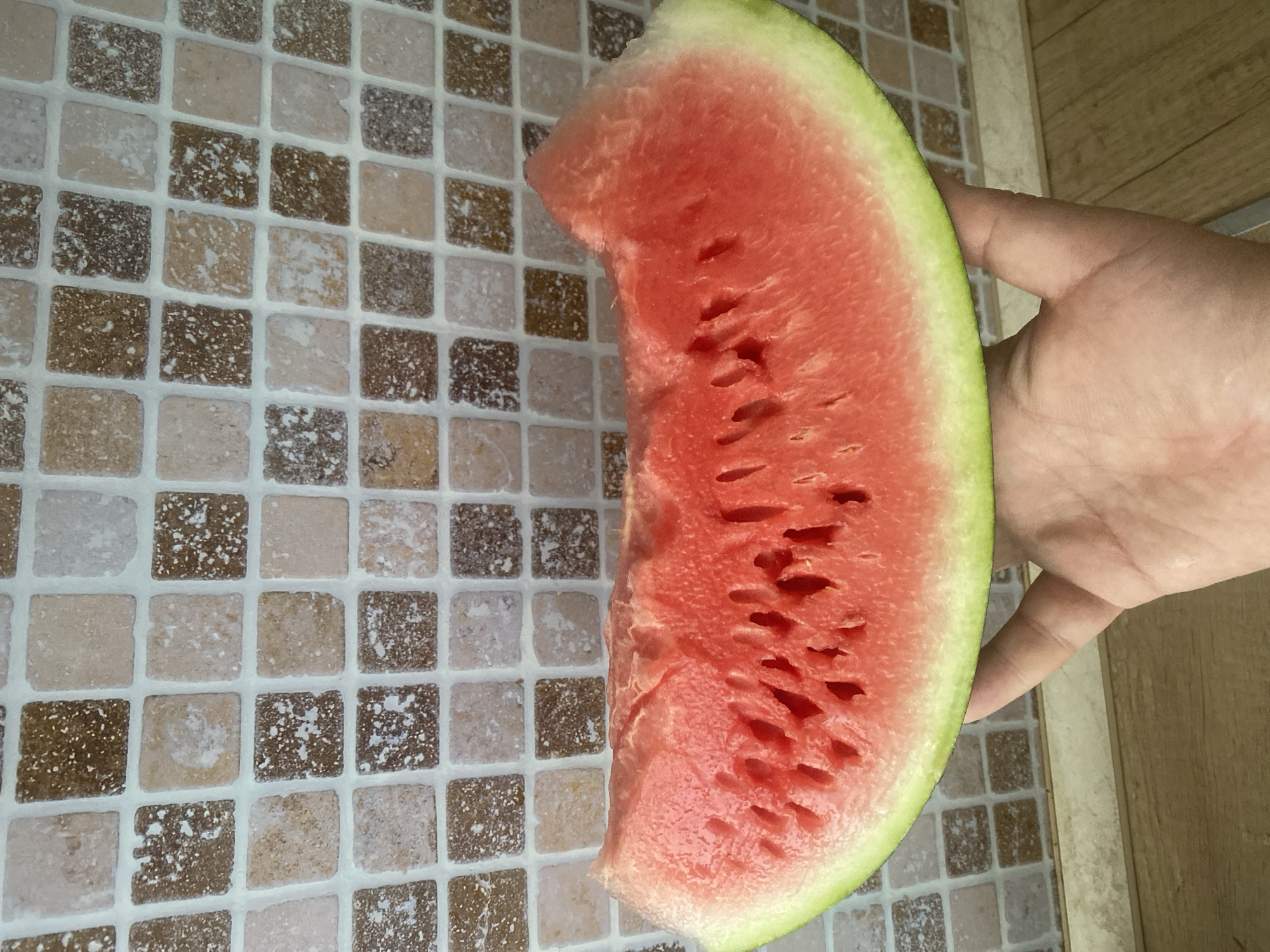 A slice of watermelon is being held by a person's hand against a tiled background. The watermelon is bright red with some seeds visible, and a green rind. The tiles behind it are small and square, with a mottled brown and beige pattern. The background suggests an indoor setting, possibly a kitchen or bathroom. There is no indication of the location being in Chișinău, Moldova.