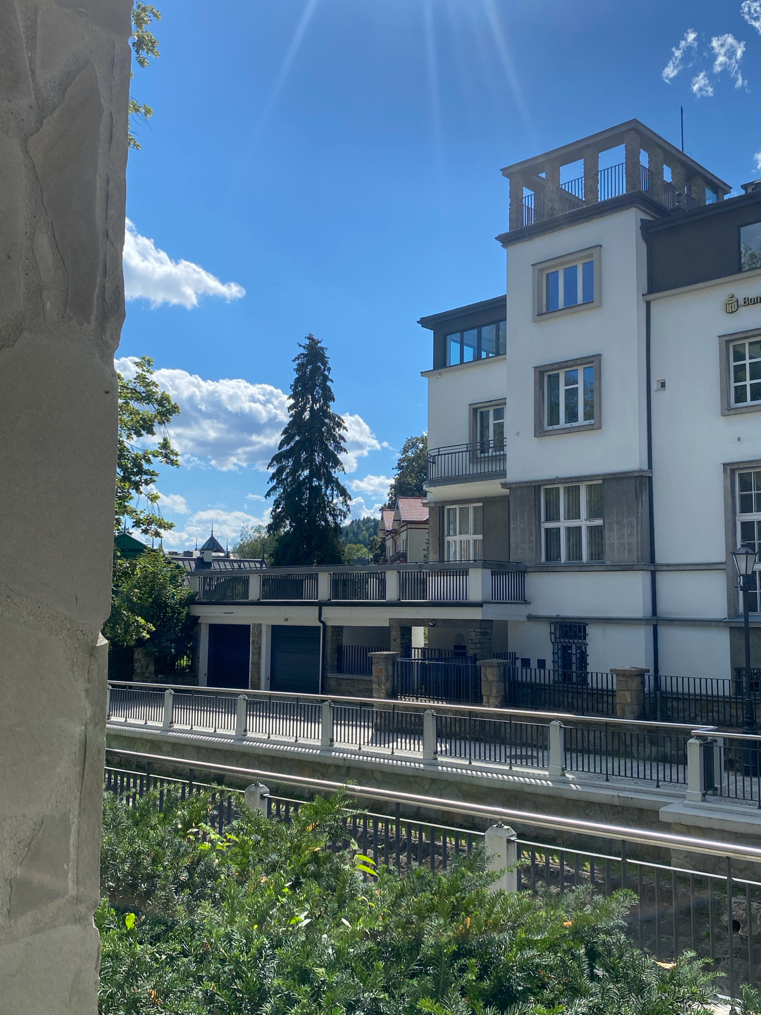 The image depicts a view of a building in Krynica-Zdrój, Poland, likely a sanatorium or hotel, with a focus on its architecture and surrounding environment. The building is a multi-story white structure with dark-framed windows, balconies, and a rooftop terrace with stone columns. In the foreground, a large, rough-textured stone structure is visible on the left, partially obscuring the view. A metal railing with white pillars runs horizontally across the mid-ground, separating a walkway from a lower level with garages. Lush green foliage fills the bottom of the frame. In the background, a tall, slender evergreen tree stands prominently, with other trees and buildings, including houses with red roofs, visible further in the distance. The sky is a vibrant blue with scattered white clouds, and sunlight streams down from the upper left, creating lens flare effects. No people are present in the image.