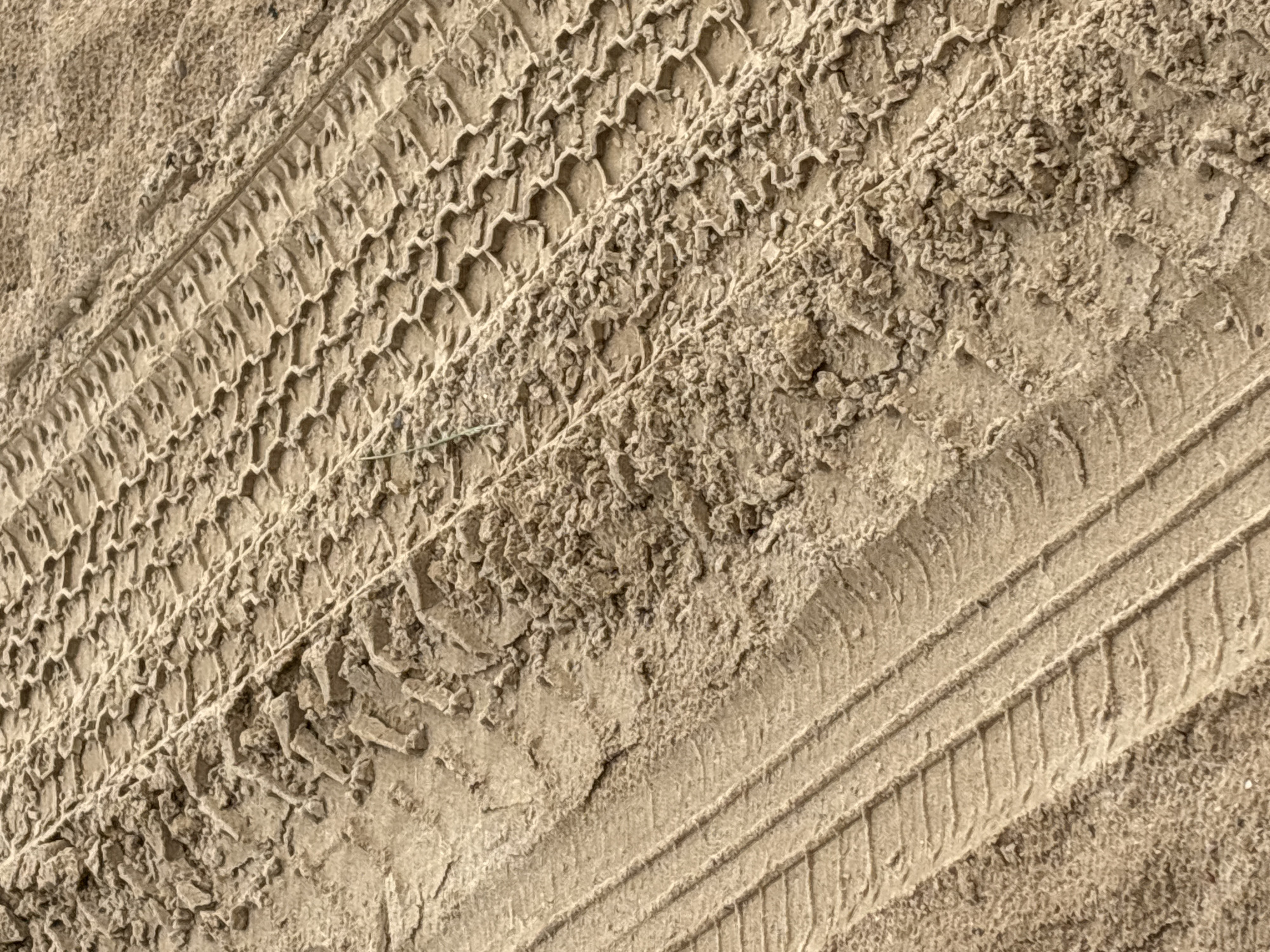 The image shows tire tracks in sandy soil. The tracks are deep and distinct, indicating that a heavy vehicle has recently passed through. The sandy soil appears to be somewhat damp, as evidenced by the way the sand has clumped and formed ridges within the tire impressions. There is a hint of green vegetation in the upper right portion of the image, suggesting an outdoor environment. The overall impression is of a rural or undeveloped area, possibly a construction site or a dirt road. The location is specified as Ждановіцкі сельскі Савет, Belarus. There are no people or animals visible in the image.