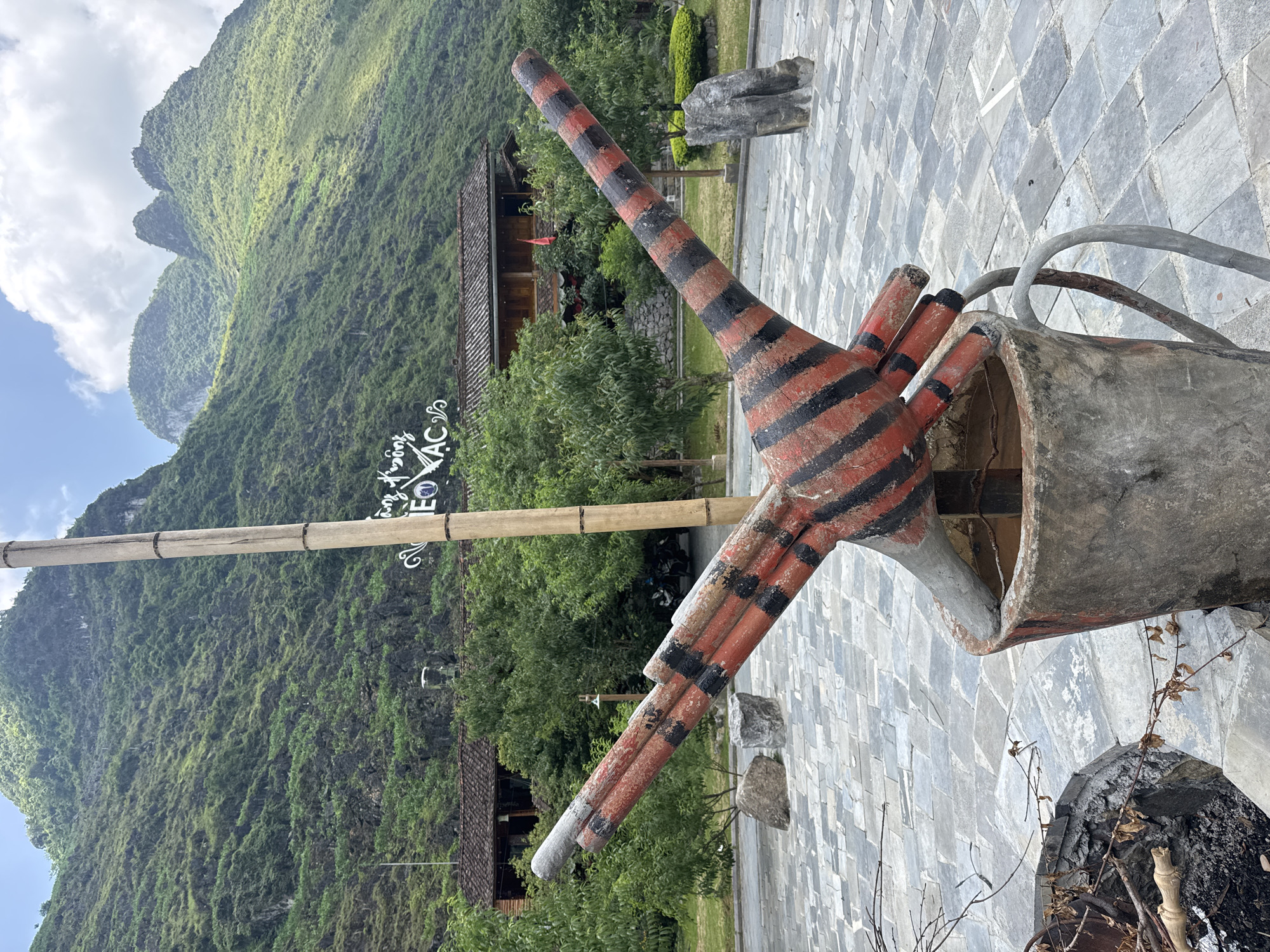 The image shows a rural scene in Mèo Vạc Commune, Vietnam, featuring a large, abstract sculpture of a watering can with multiple spouts, painted with red and black stripes. The can appears to be made of concrete. It is set on a paved area. In the background, a lush, green mountainside dominates the landscape under a sky with light clouds. A bamboo structure with a sign in Vietnamese is also visible. The surroundings include vegetation, a building with a tiled roof, and scattered rocks.