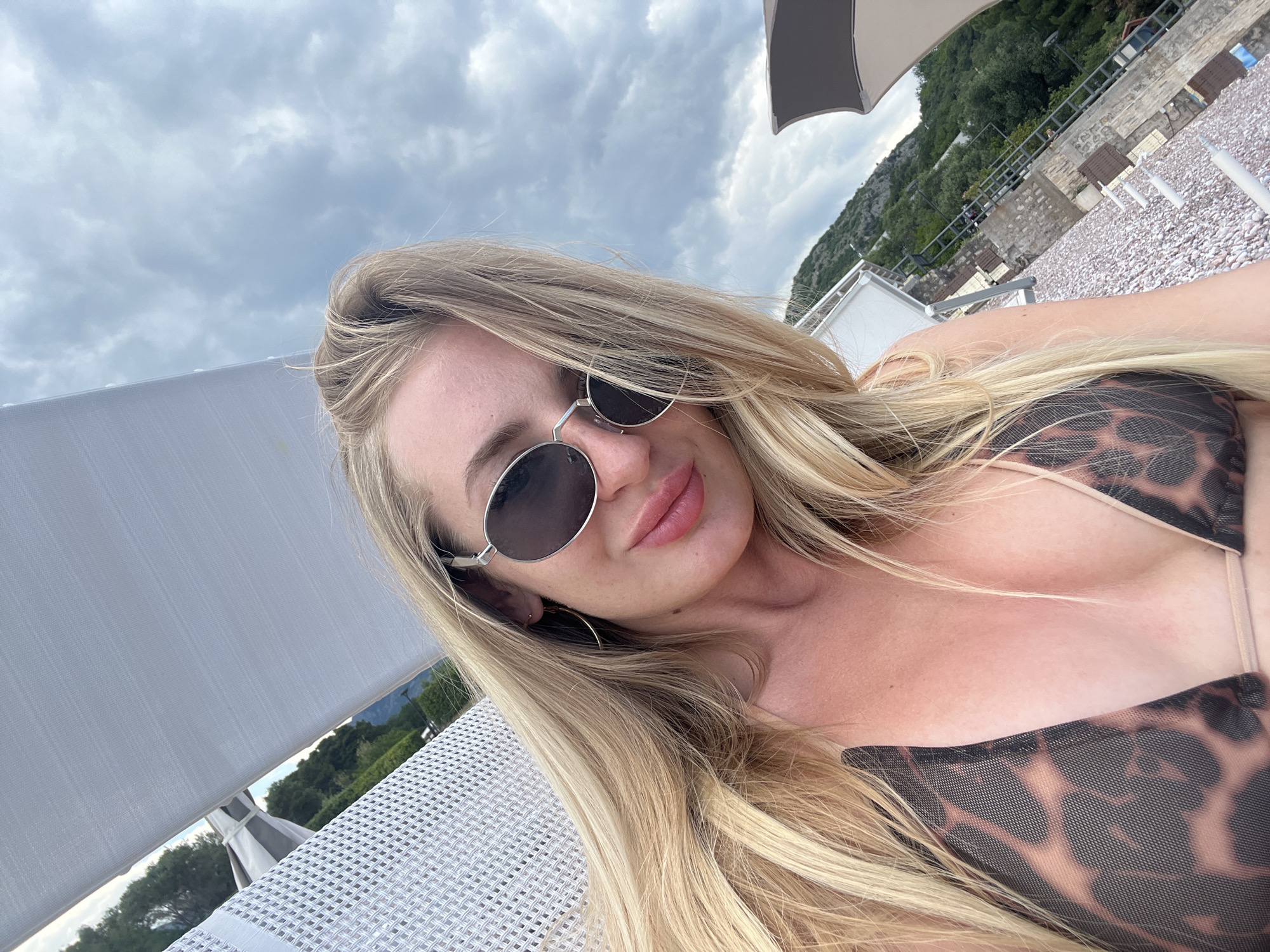 A blonde woman wearing round sunglasses and a leopard print bikini lies on a white mesh sun lounger on the beach in Sveti Stefan, Montenegro. Overcast skies fill the top of the frame, with mountains visible behind her. A beach with pebbles and additional sun loungers and umbrellas are partially visible in the background.