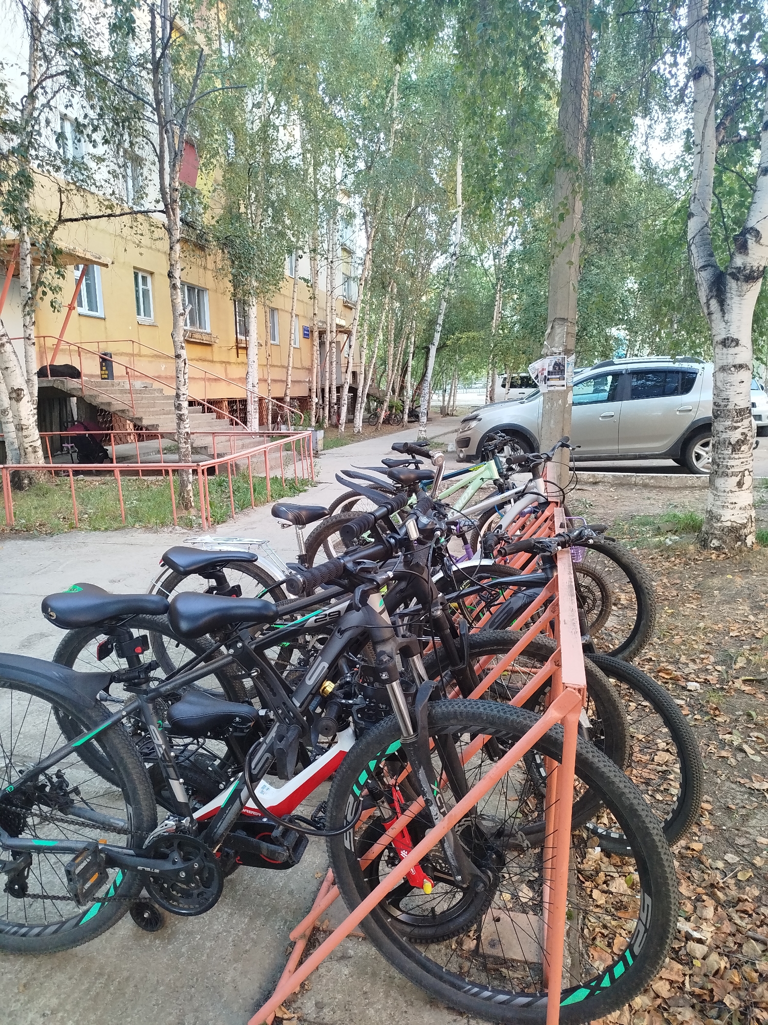This image depicts a row of parked bicycles in front of a yellow apartment building in the city of Mirny, Russia. The bicycles are primarily mountain bikes, with various frame colors including black, white, and light green. They are secured to a coral-colored bike rack. The ground is paved with concrete and scattered with fallen leaves, suggesting an autumn season. Trees with white bark and green leaves line the area, with some trees appearing to be birch. In the background, a silver SUV is parked near the building, and a staircase with a red railing leads up to the entrance of the apartment building. The overall scene is calm and orderly, with no people visible in the frame.
