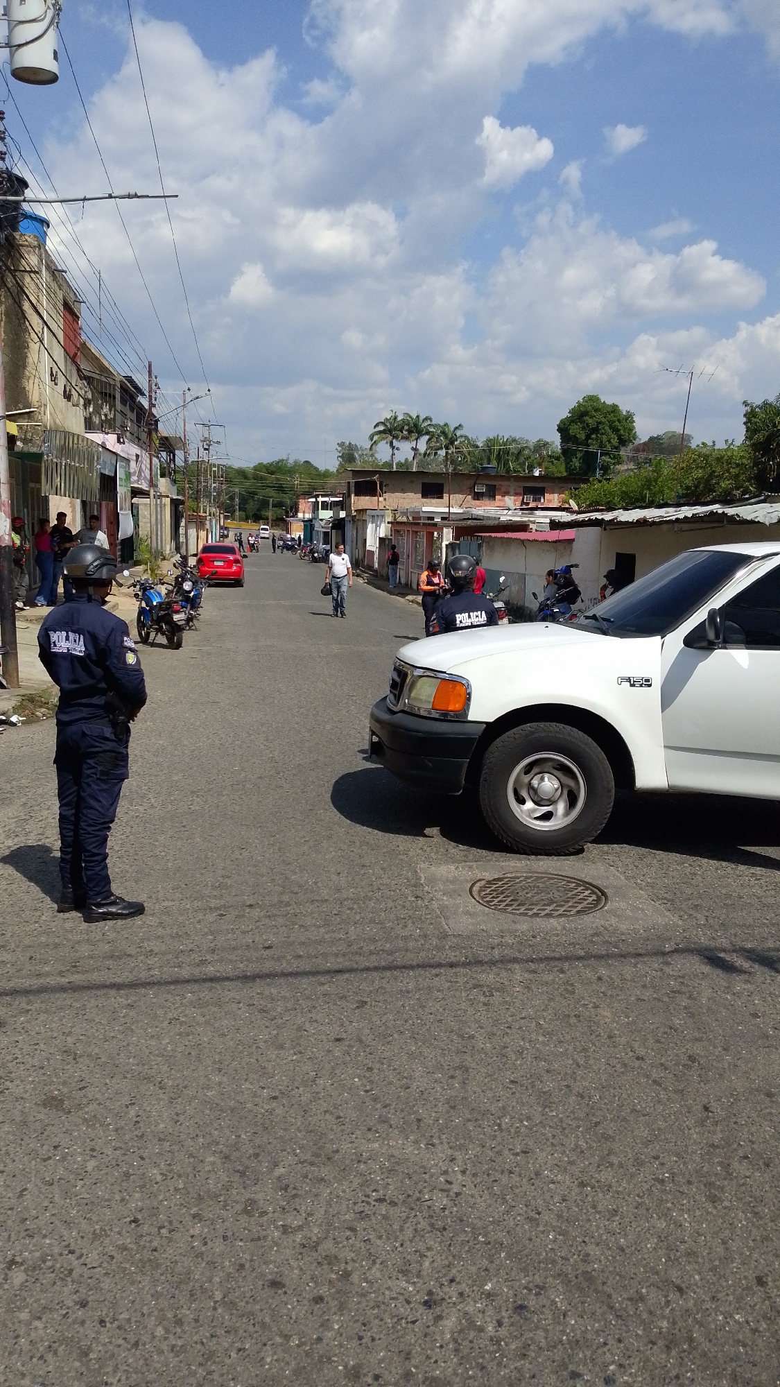 The image depicts a street scene in Tinaquillo, Venezuela. A police officer in a dark blue uniform and helmet stands on the left side of the road, facing forward. A white Ford F-150 pickup truck is parked on the right side of the road, partially obstructing the view down the street. Another officer, similarly dressed, stands near the vehicle. A red car is visible further down the street along with motorcycles parked on the side of the road. Pedestrians walk in the distance. Buildings line both sides of the street. The sky is partly cloudy. A manhole cover is visible on the road in front of the truck. Power lines are strung overhead.