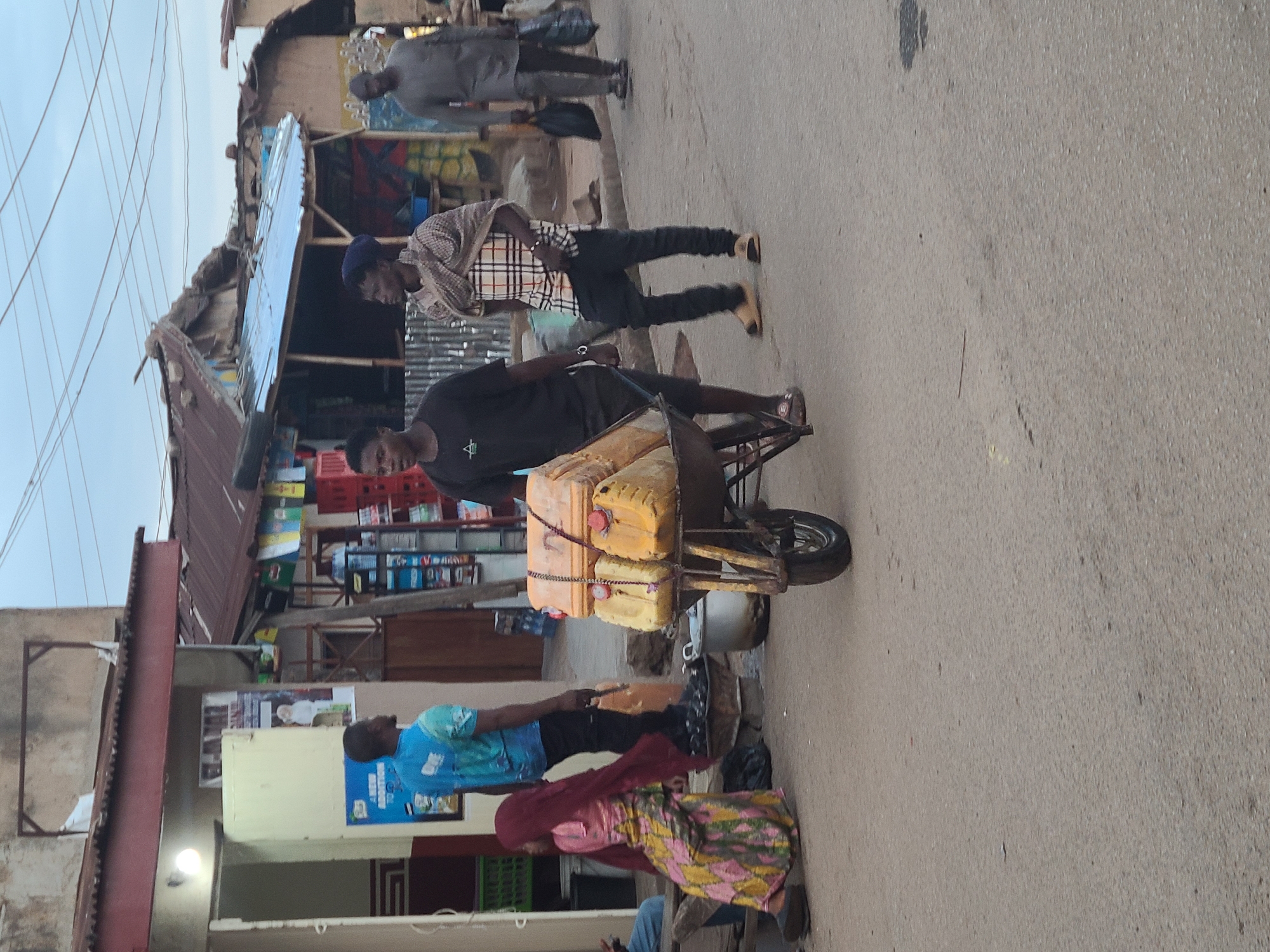 The image depicts an outdoor street scene, likely a bustling market or commercial area, with several people engaged in daily activities. The setting appears to be an urban or semi-urban environment, characterized by rustic buildings with corrugated metal roofs and walls, and overhead electrical wires crisscrossing the sky.
In the foreground and central to the image, a man wearing a black t-shirt with a white triangular logo is pushing a makeshift cart or wheelbarrow. The cart is heavily laden with several yellow jerrycans, suggesting he might be transporting water or other liquids for sale or delivery. A metal pot or bowl rests beneath the cart. Behind him, another man stands, dressed in a plaid long-sleeved shirt and dark trousers. Further back, a man in a grey long-sleeved outfit stands.
To the left, a man in a blue patterned t-shirt is visible next to a building. Beside him, a woman in a colorful patterned skirt and a red headscarf is either sitting or crouching low. The background features various stalls or shops, evidenced by stacks of red crates and other merchandise. Posters and signs are affixed to the building walls; one blue sign prominently displays the partial text "NEW ADOPTION" and other illegible words below it. Another light fixture is visible on a building facade.
The ground is a paved street or pathway, showing some wear and minor debris. The sky is overcast, providing soft, diffused daylight without harsh shadows, suggesting either an overcast day or early morning/late afternoon. The overall scene captures a slice of everyday life, focusing on people going about their business in a lively street setting.