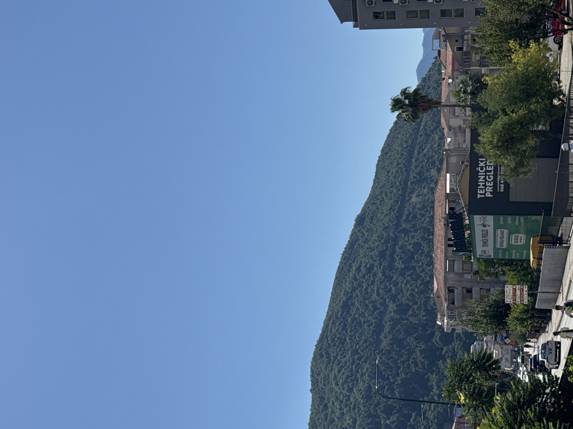 This is a photograph of a street scene in Budva, Montenegro, taken on a clear, sunny day. The image is oriented vertically, with the sky occupying the left half of the frame and a steep, tree-covered mountain rising on the right. Buildings line the street at the base of the mountain, interspersed with trees, including a prominent palm tree.
On the right side of the street, a building displays a large green sign with white text that reads "TEHNIČKI PREGLED" and "SAIA TEHNIČKI PREGLED MÄINE." Below this, there is additional text, possibly indicating a phone number or address. Further down the street, several cars are visible, suggesting moderate traffic. People can be seen walking on the sidewalk. The time of day appears to be late morning or early afternoon, given the bright sunlight and clear shadows. The overall impression is one of a typical urban environment nestled against a dramatic natural landscape.