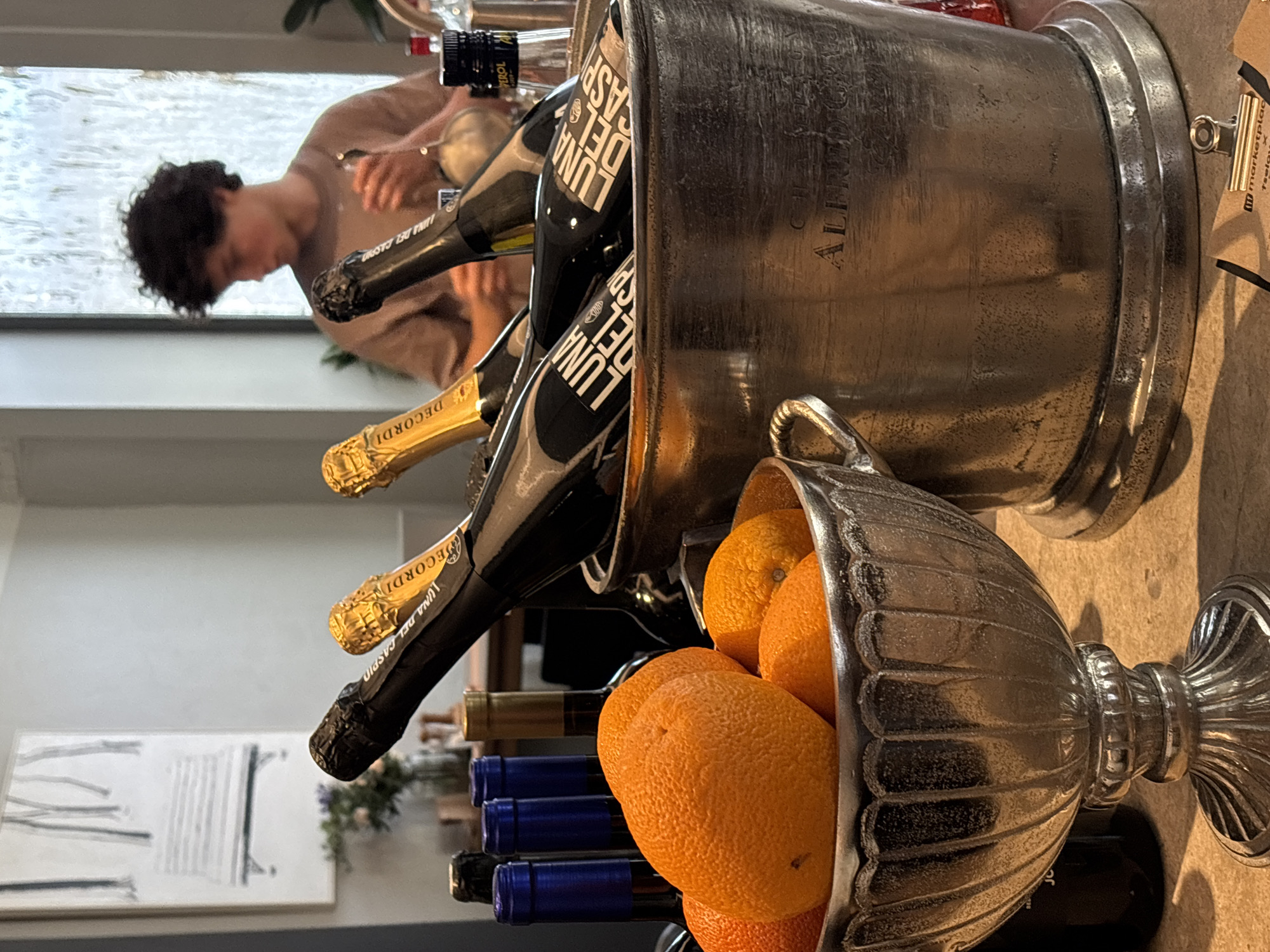 This image shows a close-up of a metallic ice bucket filled with bottles of sparkling wine. The bottles have labels with "LUNA DEL" and "CASA" visible. In front of the ice bucket, a silver urn-shaped bowl is filled with oranges. In the background, a young person with curly hair is visible, wearing a light brown t-shirt, looking down and holding a small object in their hands, possibly preparing a drink. Behind the person, a window with textured glass or water feature is visible. To the left of the scene, there are other bottles, some with blue caps, and a framed artwork on the wall depicting minimalist trees. The setting appears to be a bar or restaurant, likely in Saint Petersburg, Russia, given the context of the image's origin. The lighting is soft, suggesting an indoor environment.