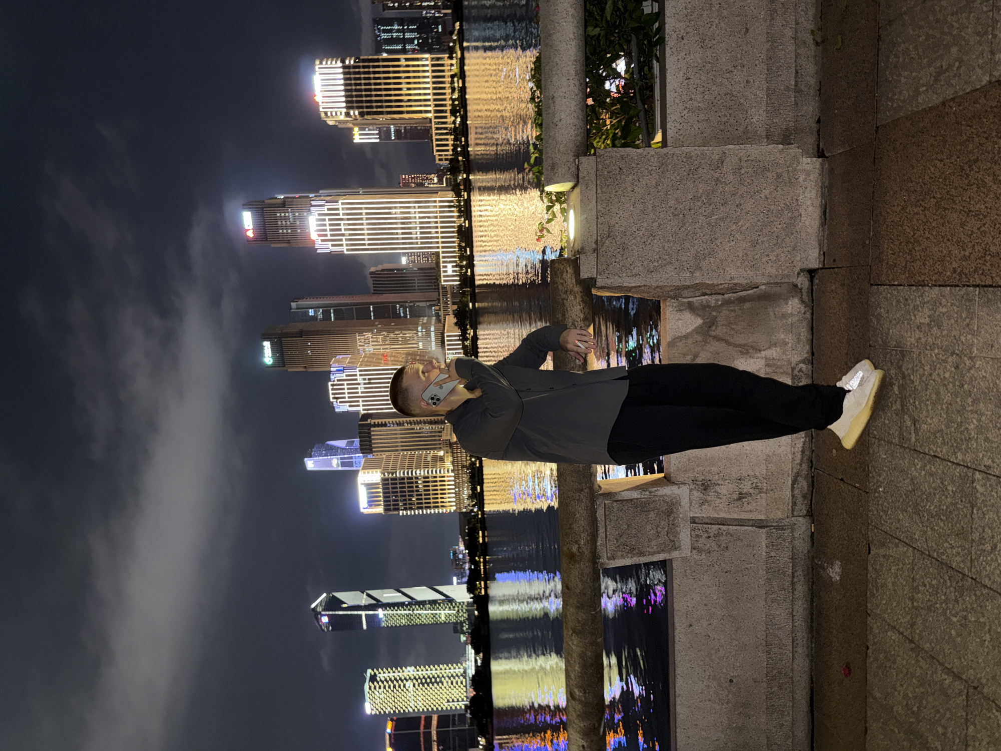 The image captures a nighttime urban scene along a waterfront in Tianhe District, China. A male adult, fair-skinned with short light-colored hair, is positioned in the foreground. He wears a dark grey long-sleeved collared shirt, black trousers, and light-colored athletic shoes with reflective elements on the heels. He stands with his left arm resting on a textured stone barrier, holding a light-colored smartphone to his right ear, appearing to be engaged in a call.
The immediate foreground features a paved walkway composed of large, rectangular stone tiles. This leads to the rough-hewn stone barrier, which includes an integrated light fixture on its top surface emitting a warm yellow glow. Some green foliage is visible behind the barrier on the right side.
Beyond the barrier lies a wide river or canal, reflecting the vibrant, multi-colored lights from the distant cityscape, predominantly yellow, blue, and purple.
The background is dominated by a dense array of modern high-rise buildings across the water, illuminated against a dark, overcast sky. The skyscrapers feature prominent architectural lighting, primarily vertical strips of warm white and yellow light. Distinct structures include a tall building on the center-left with a striking black and white diagonal pattern, and several other towers displaying corporate logos, such as a red logo on one and a green logo on another, at their upper levels. The sky above is dark and heavily covered with grey clouds.