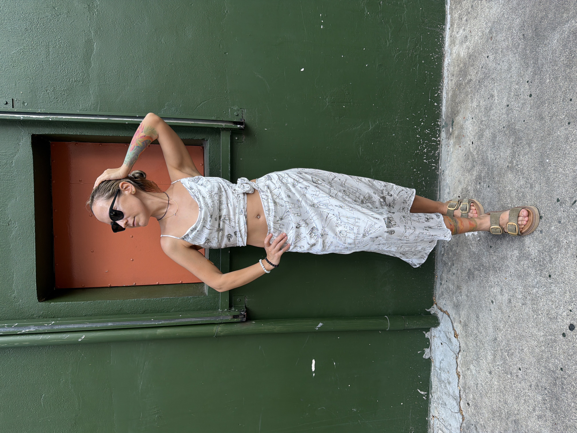 A woman is standing against a green wall with an orange panel above her. She is wearing a white patterned top and skirt, sunglasses, and sandals. Her left arm is raised, touching her head, and her right hand is on her hip. The ground is concrete, and there is a pipe on the left side of the image.