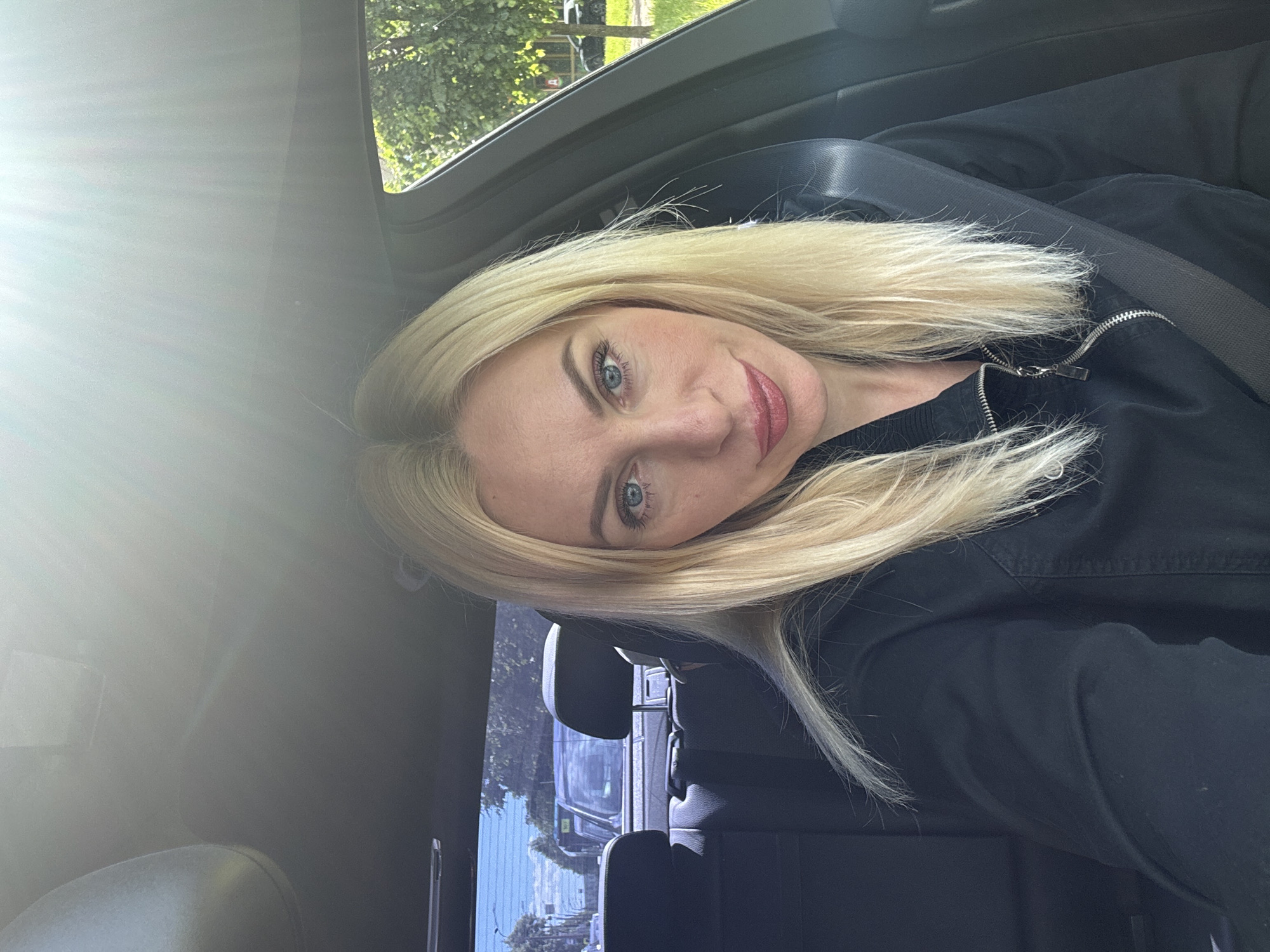 A fair-skinned blonde woman with blue eyes and a light pink lipstick is sitting in the driver's seat of a car, looking at the camera. She is wearing a black zippered jacket with a seatbelt across her chest. The interior of the car is visible, with black seats and a rearview mirror overhead. Outside the car, through the rear window, a street scene with trees and other vehicles can be seen. The sunlight is bright, creating lens flare at the top of the image. The location is the city of Saint Petersburg, Russia.