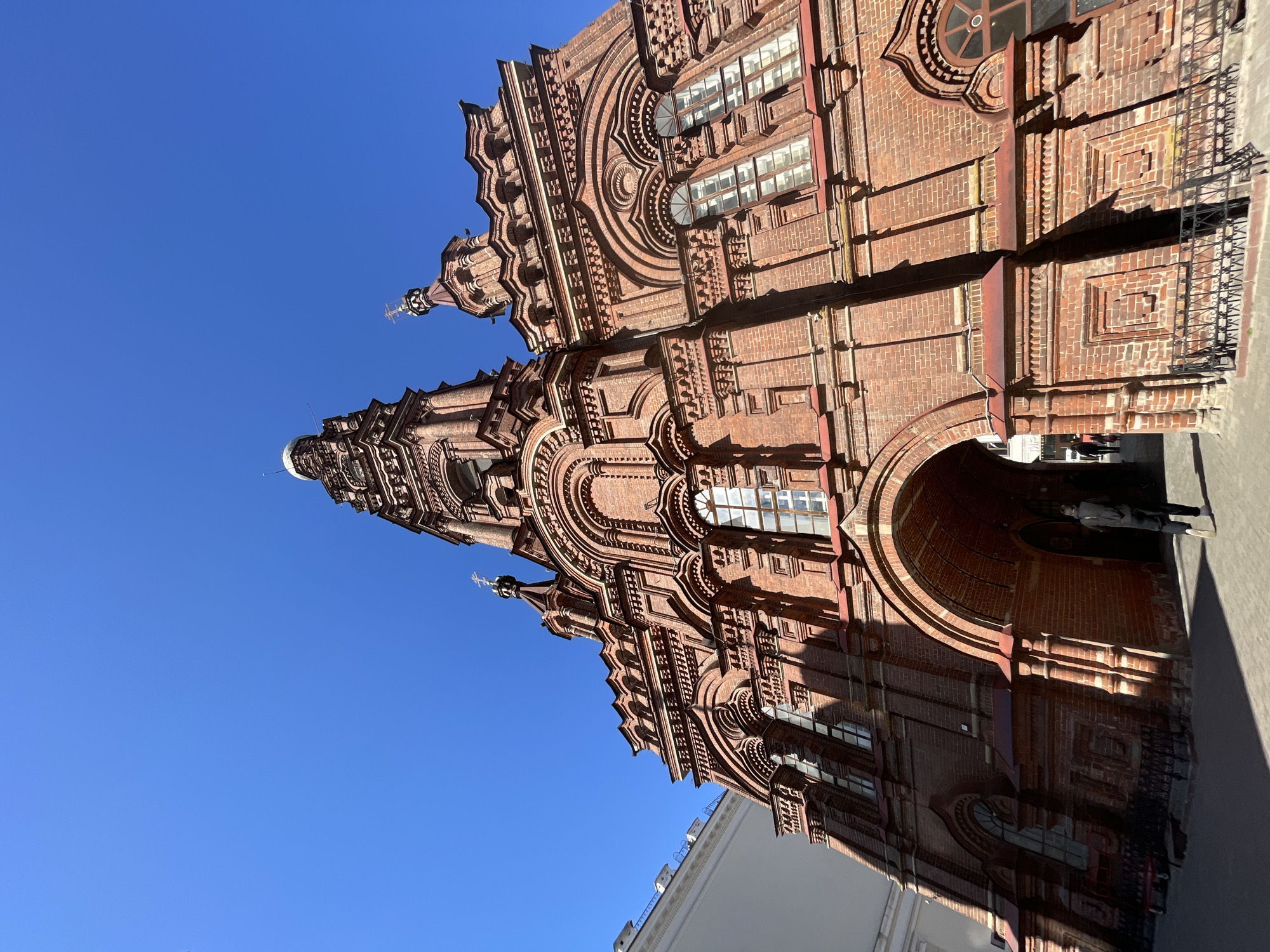The image shows a tall red brick building with intricate architectural details against a clear blue sky. The building features arched windows, decorative brickwork, and a tower. The scene is likely the Epiphany Cathedral in Kazan, Russia. The base of the building shows a large archway where a person is seen walking through. A shadow of a nearby building is cast upon the right side of the frame. Part of a white building can be seen in the lower portion of the frame. The photo is oriented so the building appears diagonally across the frame, extending from the lower right to the upper left.