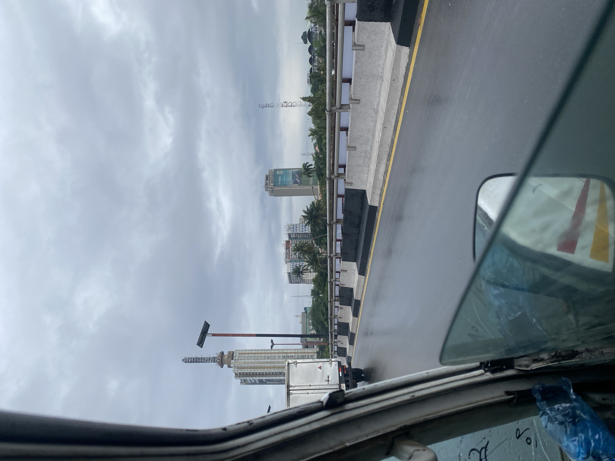 The image depicts a view from inside a vehicle traveling on a wet road, likely a bridge, in Victoria Island, Nigeria. The sky is overcast with clouds. In the background, several tall buildings and structures are visible, including a distinctive tower with a communication antenna on top, and other skyscrapers with visible signage. A white truck is in front, on the same road. The road itself is wet and reflective, indicating recent rain, with a yellow line marking the edge of the lane. Concrete barriers with black and white markings line the side of the road, separating it from the water or lower ground below, where palm trees and other greenery can be seen. The foreground shows the interior of the vehicle, including part of the windshield, side mirror, and door frame, with some plastic sheeting and writing visible on the glass.